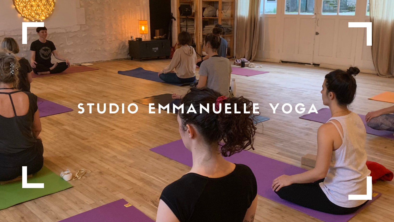Groupe de personnes participant à une séance de yoga dans un studio lumineux, assises sur des tapis de différentes couleurs, avec un instructeur assis en position de lotus au fond.