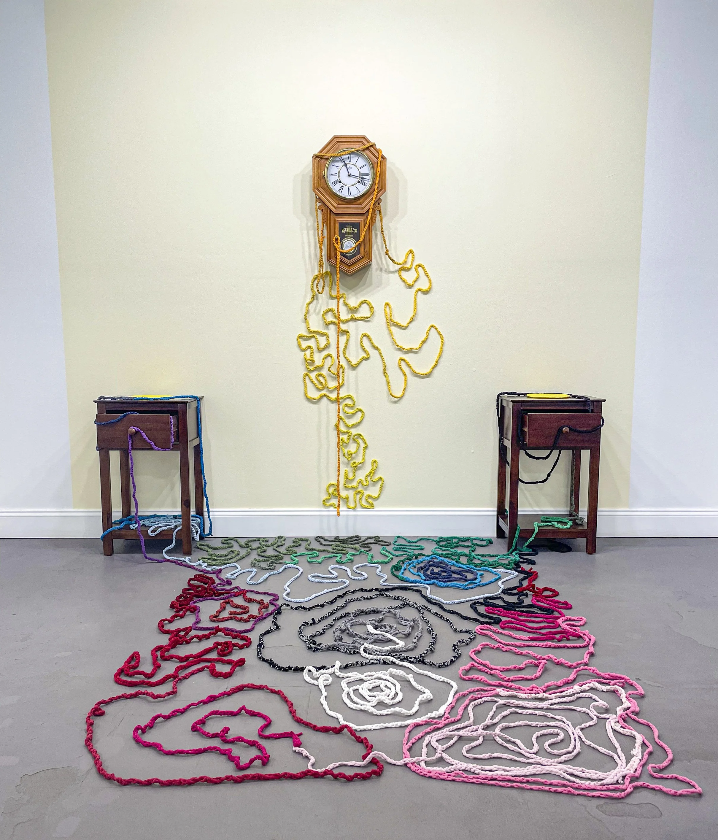  Samantha Mack,  Time and Distance,  2026. Dimensions variable, Two nightstands; two yellow trivets (one made by artist, one made by her great grandmother); threadbare hand-me-downs, cut and crocheted; chiming Regulator clock; antique book ( The Cour