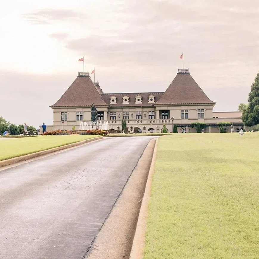 Large historic building with turrets and flags, surrounded by a green lawn and pathway, under cloudy sky.