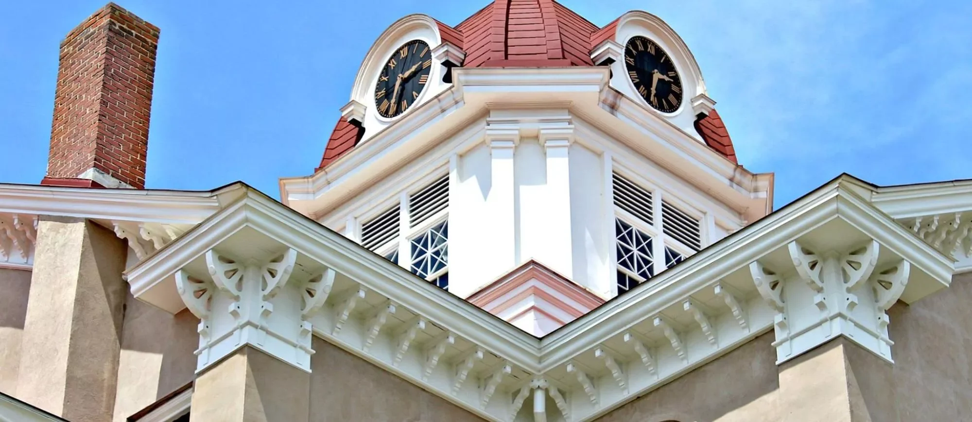 The upper part of a historic building featuring a prominent clock tower with two clocks, decorative white woodwork, a red-tiled roof, a brick chimney, and lattice-style window shutters against a clear blue sky.