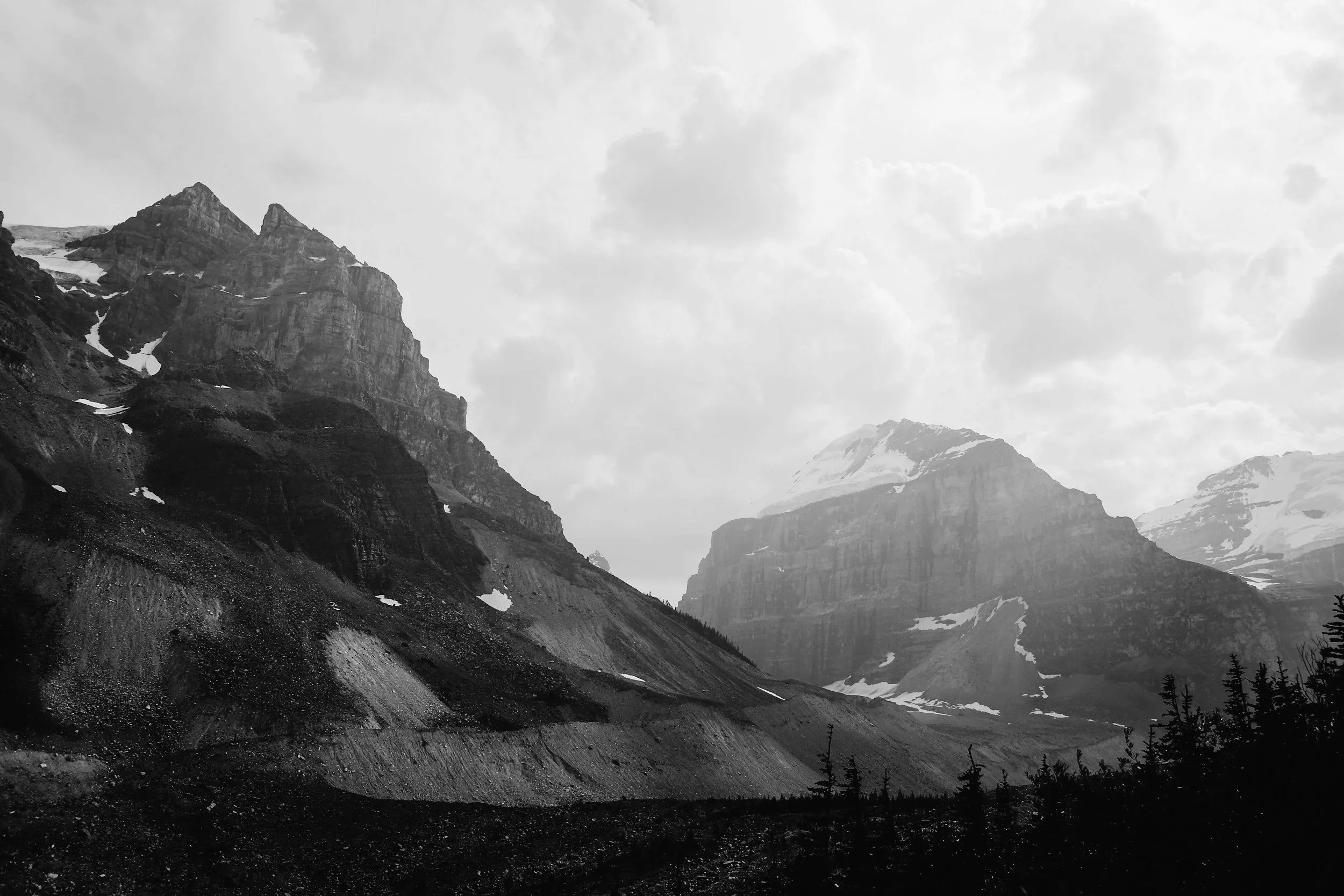 Black and white photograph of rugged mountain range with snow patches and a cloudy sky.