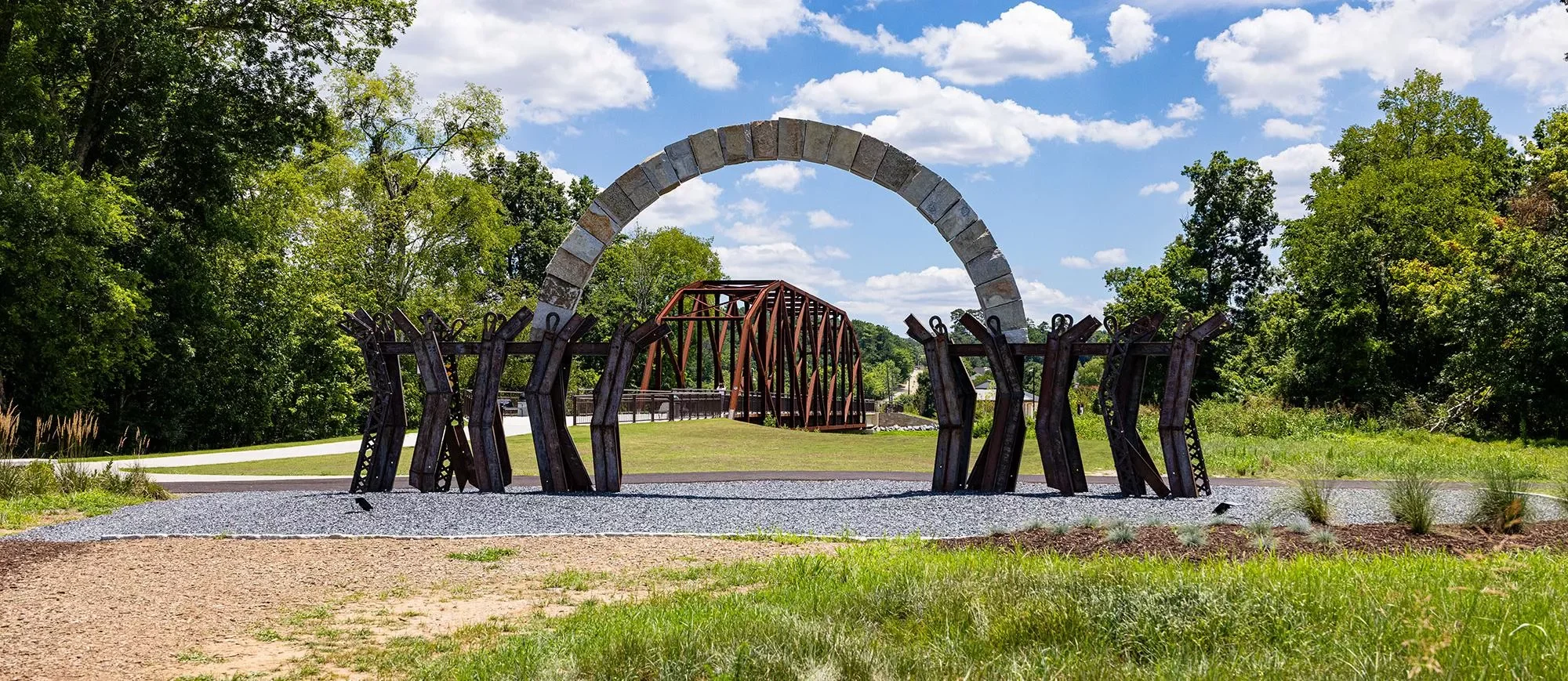 Outdoor park sculpture with metal arches, a rusty metal support structure, stone arch, and lush green trees under a partly cloudy sky.