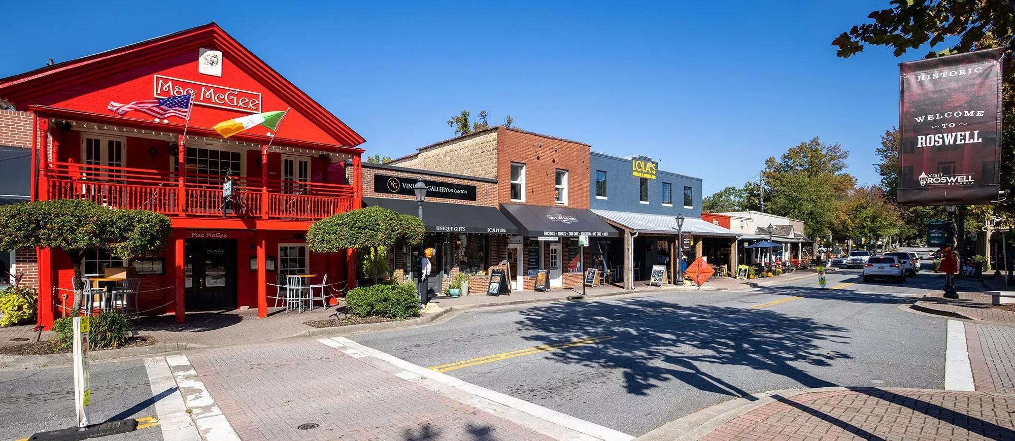 Street view in Roswell, Georgia, showing a row of small shops with storefronts and awnings, including Mac McGee restaurant with a bright red exterior, American and Irish flags, and outdoor seating. Other shops include a gallery and a store named Lola's. Cars are parked along the street, and a large welcome banner reads "Welcome to Roswell" with trees and a blue sky in the background.
