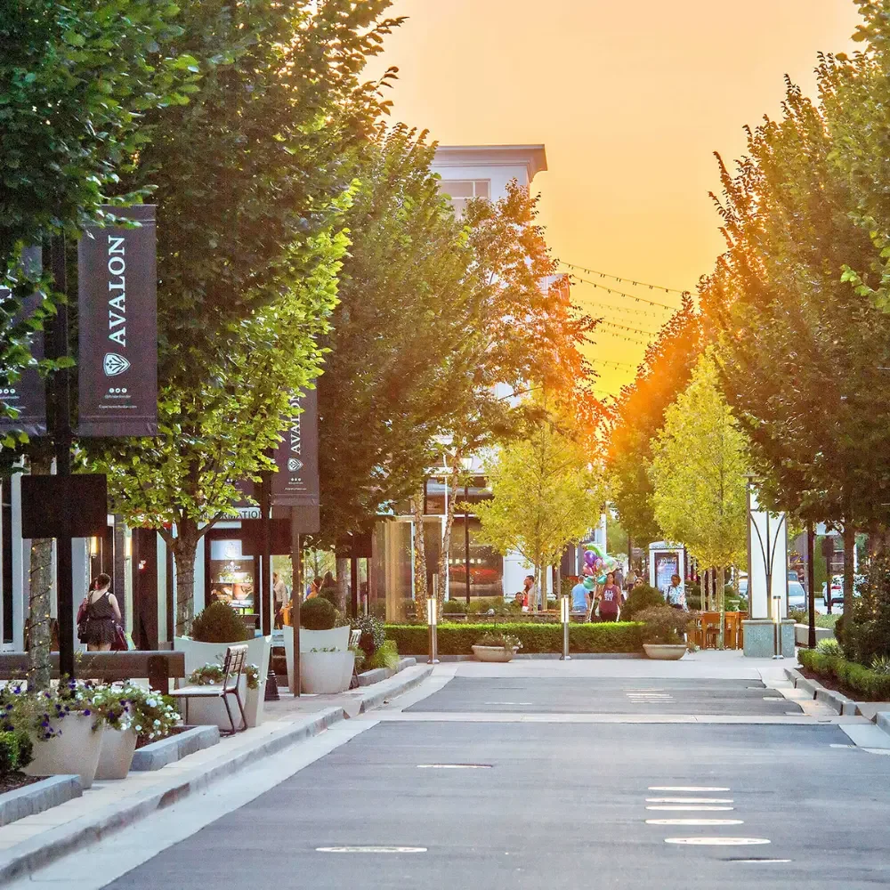 An outdoor shopping or dining street scene with trees, benches, planters, and people walking or sitting, illuminated by string lights at sunset.