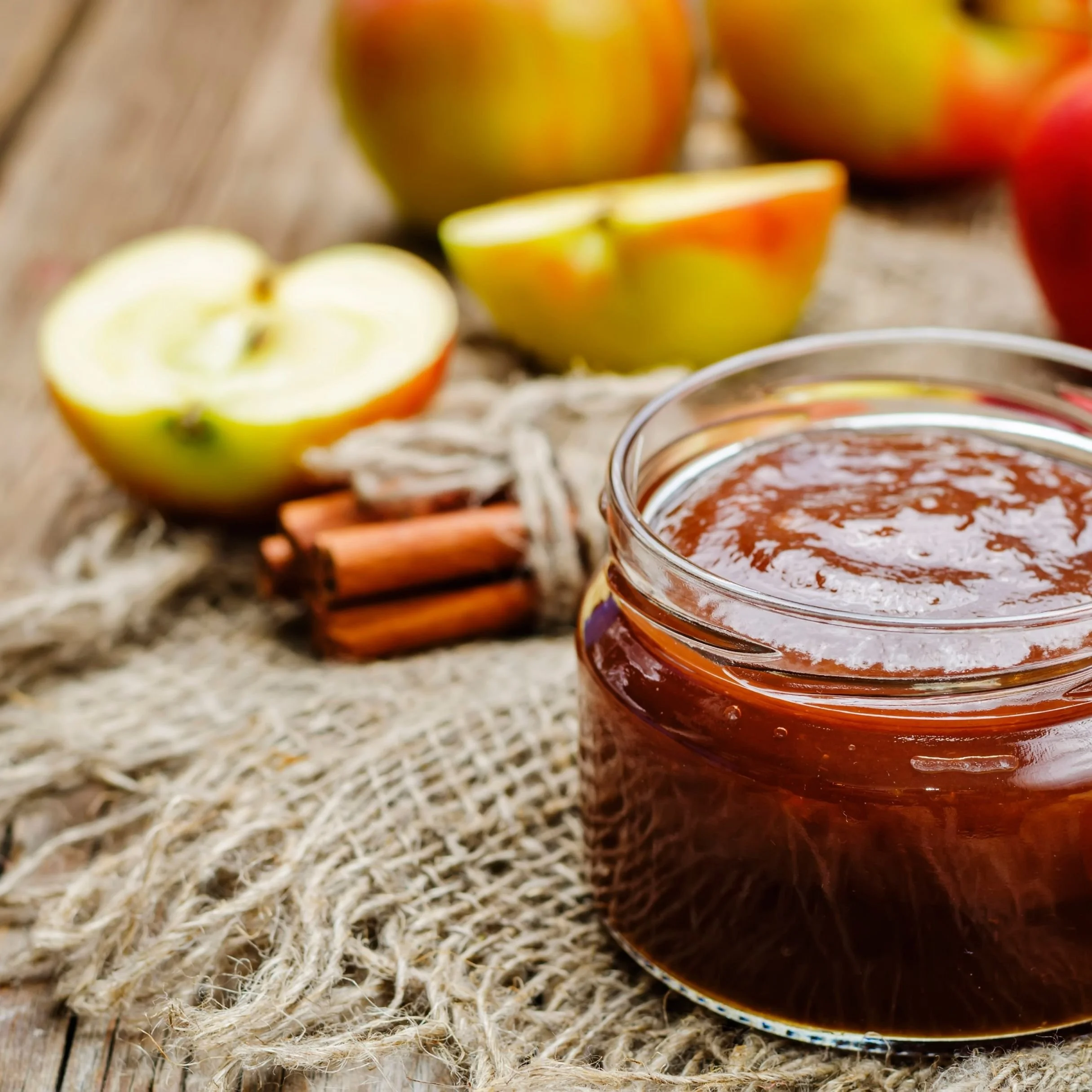 Close-up of a glass jar of apple butter on a rustic wooden surface, with halved and whole apples nearby, and pieces of cinnamon sticks.