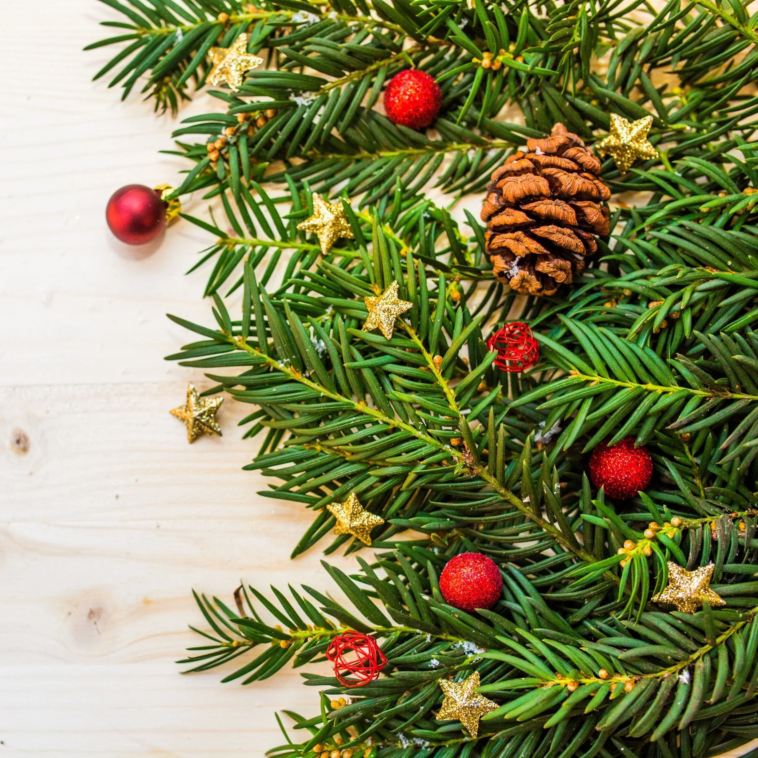 Close-up of a handmade Christmas wreath with pine branches, red and gold ornaments, pinecones, and gold star decorations on a light wooden surface.
