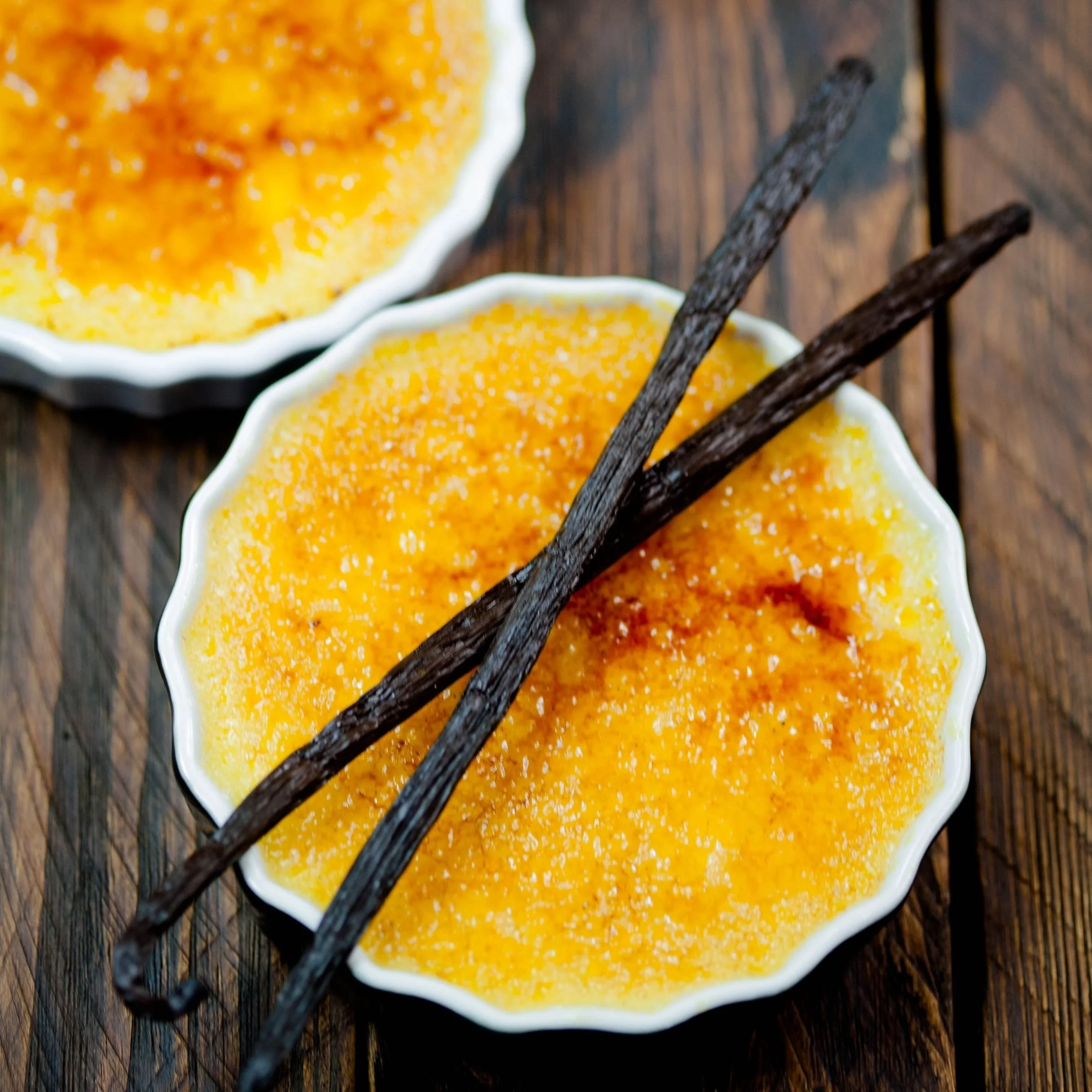 Two small white bowls filled with yellow-orange creme brulee, topped with a caramelized sugar crust, placed on a wooden surface, with two dark vanilla bean pods resting across the bowls.