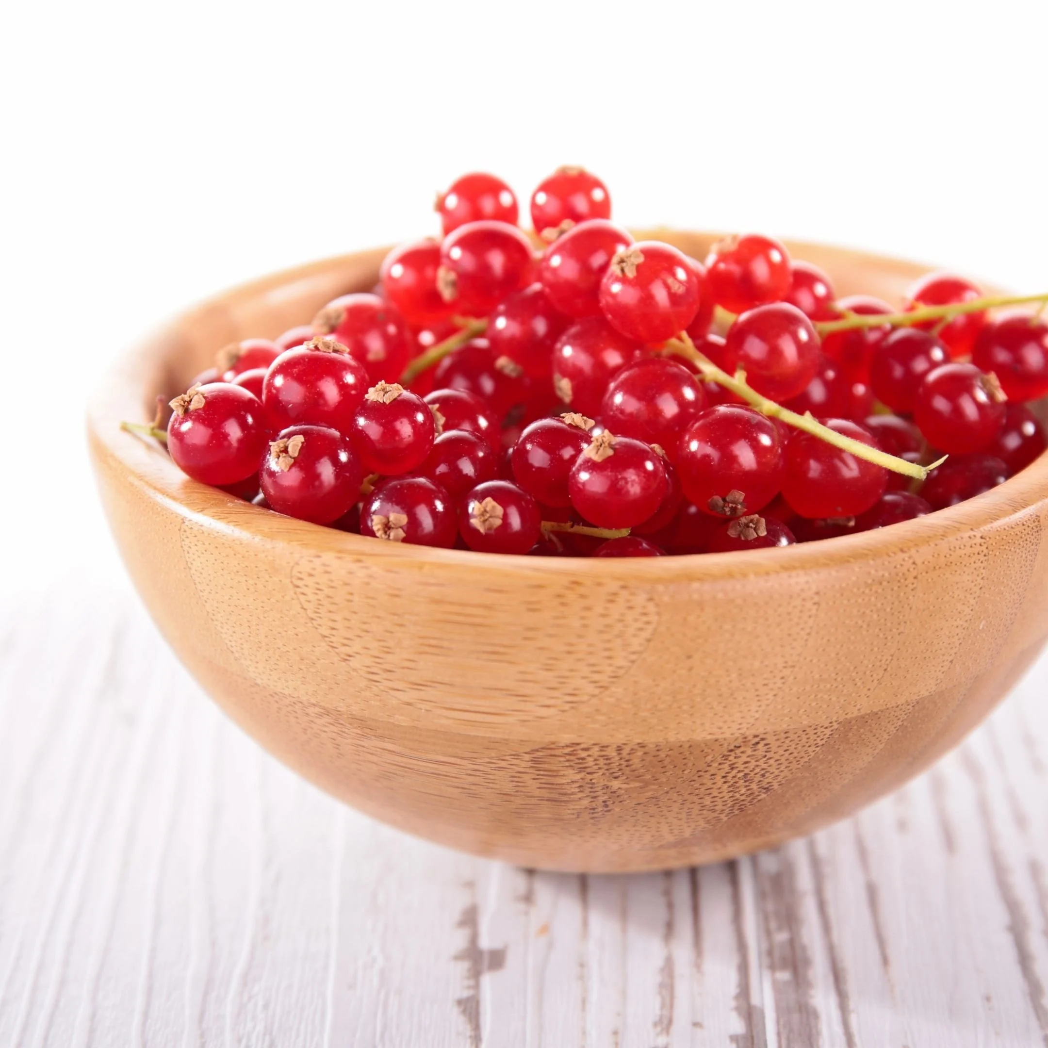 Close-up of red currants in a wooden bowl on a white surface.