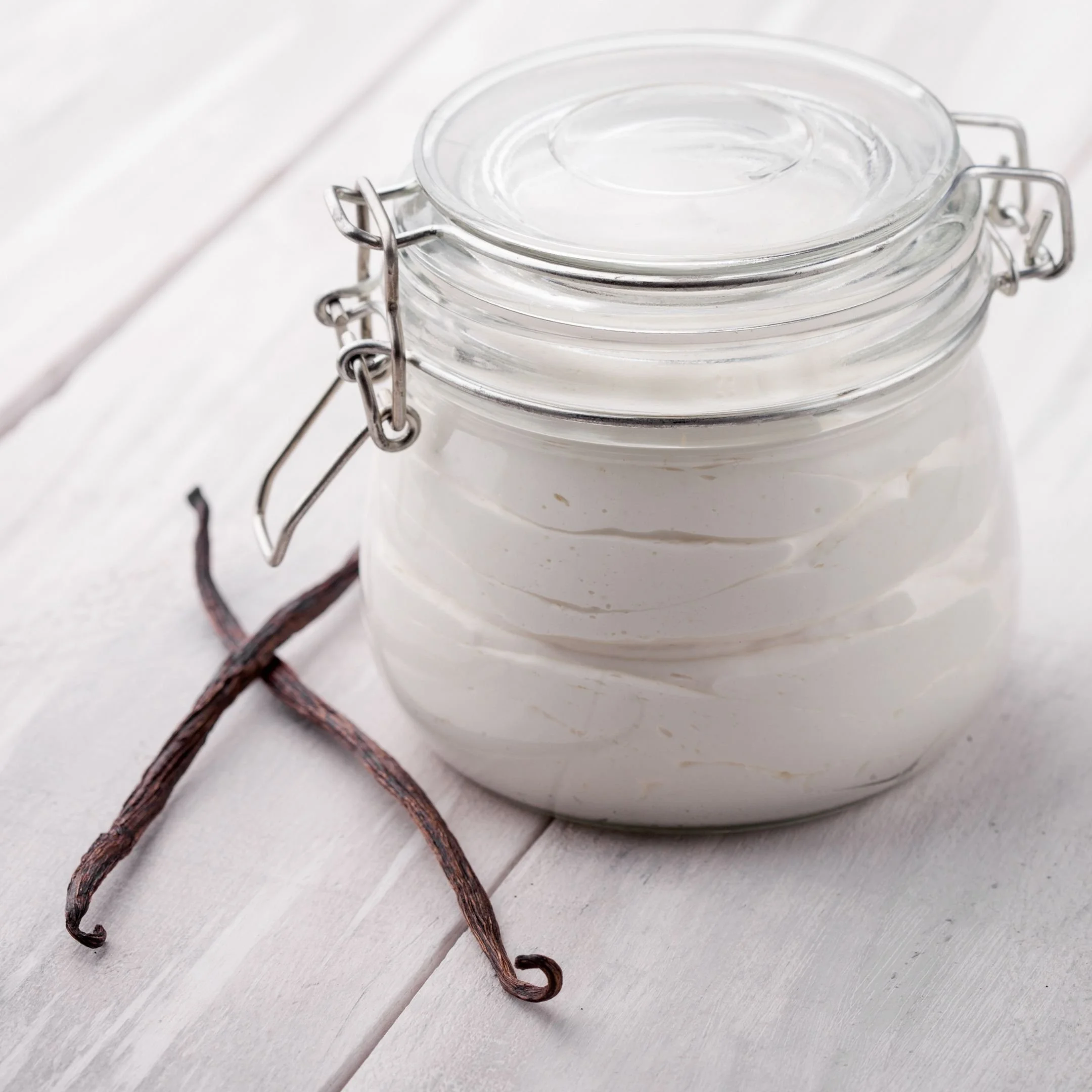 Three vanilla bean pods and a glass jar with a metal clasp lid on a white wooden surface.
