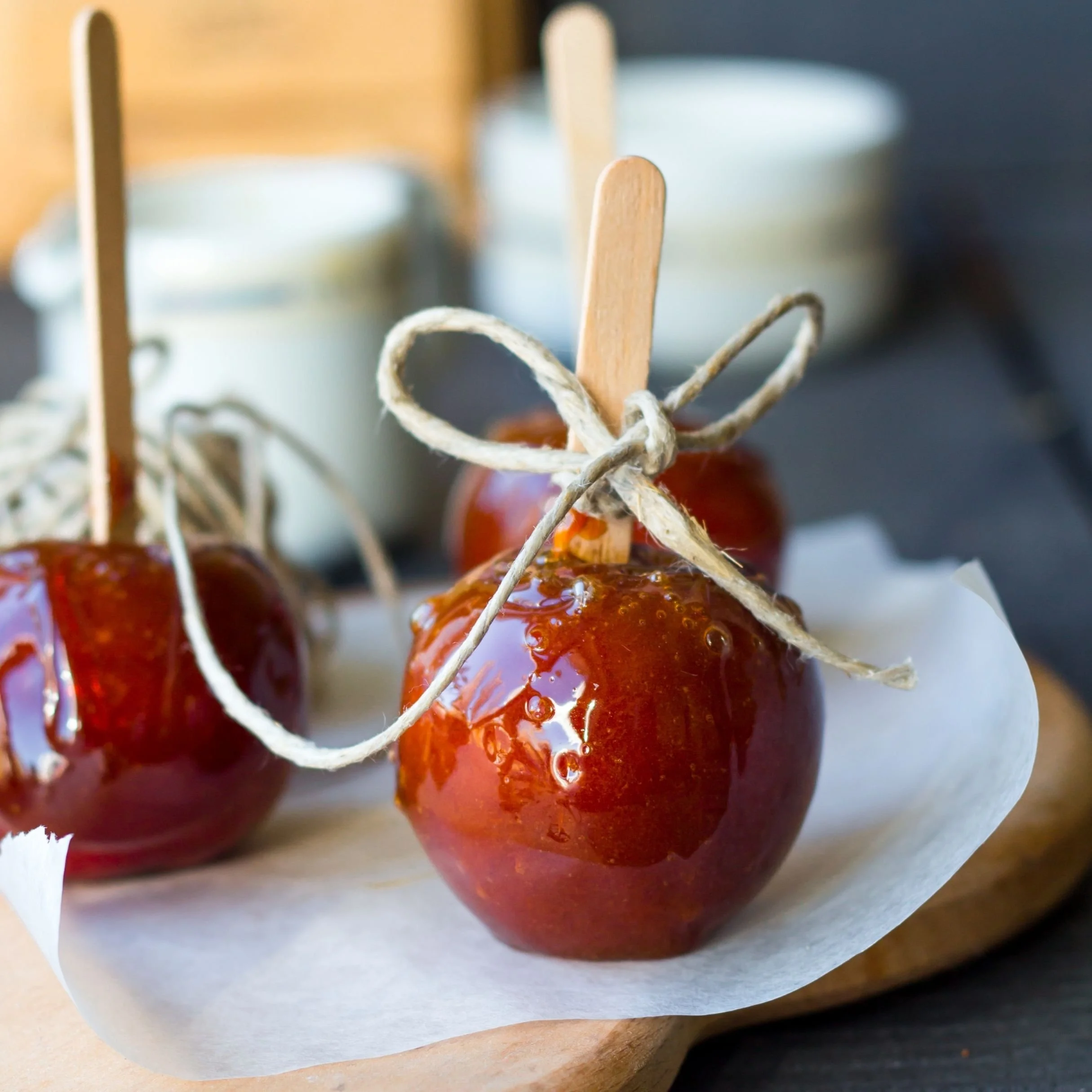 Close-up photograph of a row of glazed cherries with a wooden stick inserted in one of them, wrapped in parchment paper, on a dark background.