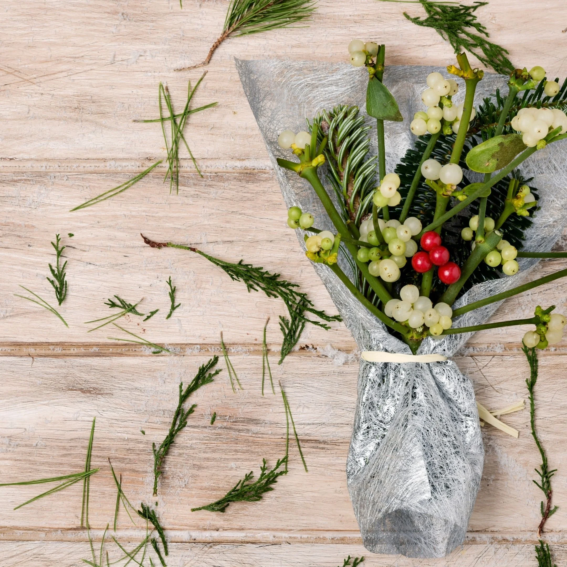 A bouquet of white berries, green leaves, and pine branches wrapped in silver mesh ribbon on a light wooden surface with scattered small pine needles.