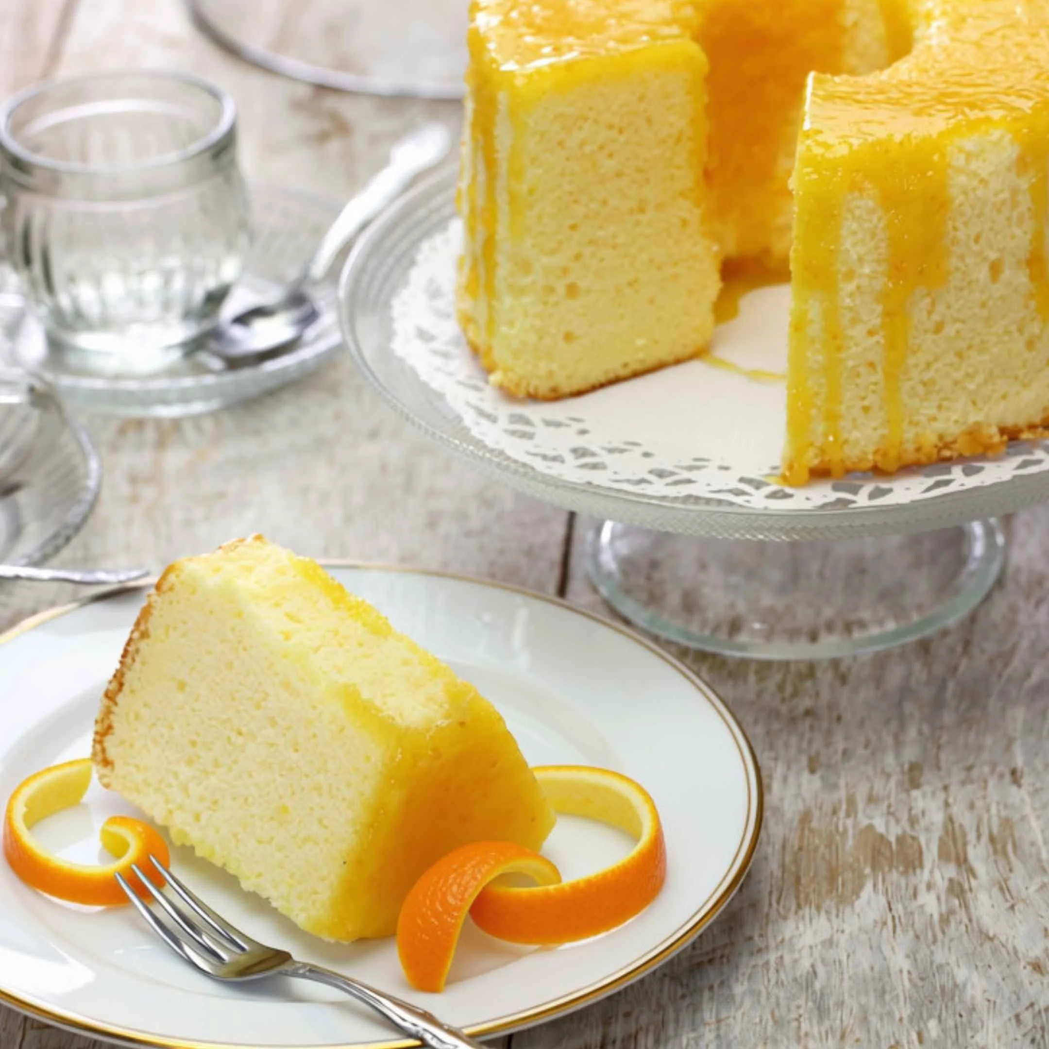 A slice of yellow cake on a white decorative plate, garnished with orange peel curls and accompanied by a silver fork, with a glass of water and a cake stand in the background.