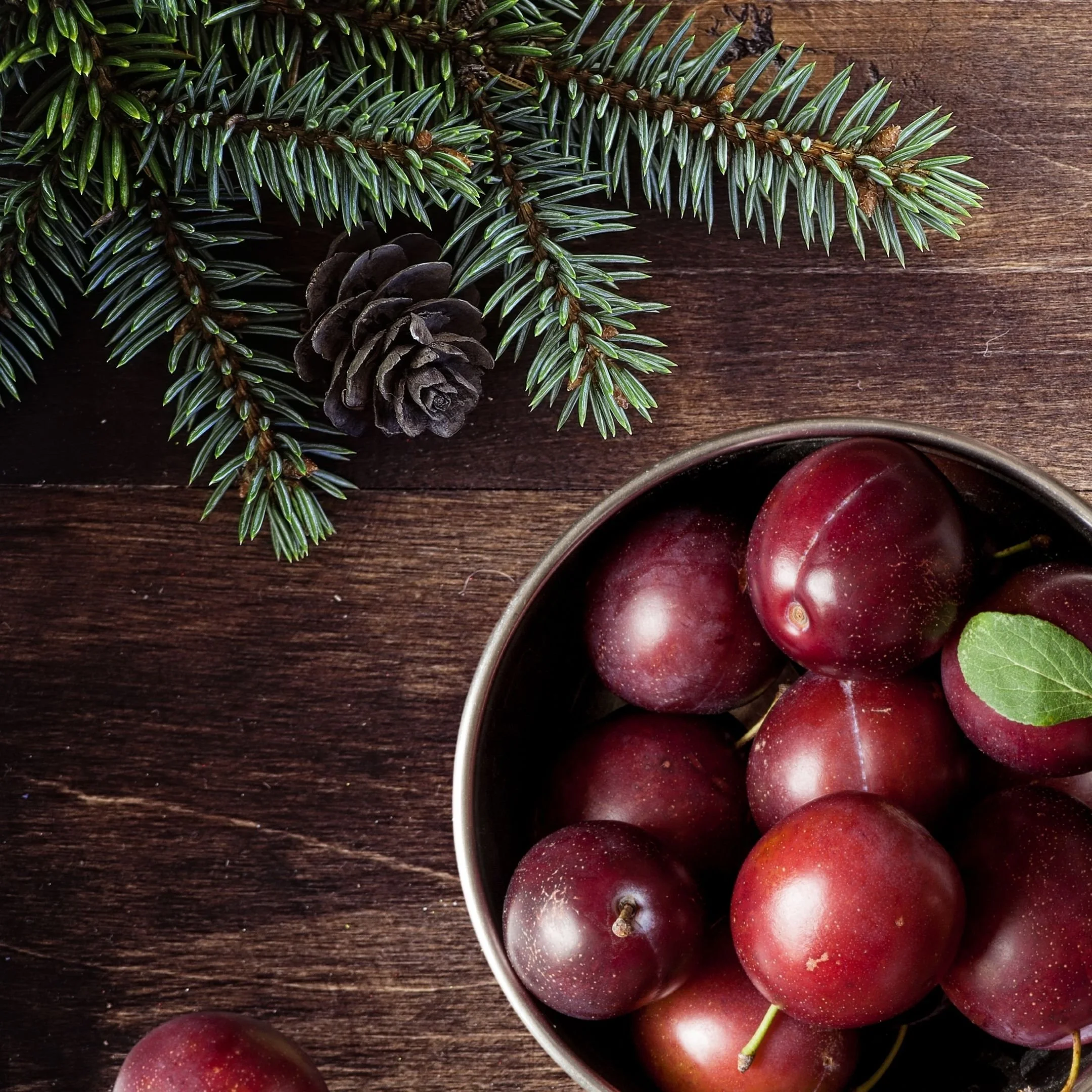 A close-up view of a bowl filled with red grapes, with some grapes and green leaves visible around the bowl on a dark wooden surface. Also included in the image are green pine branches and a pine cone, creating a rustic, festive holiday scene.