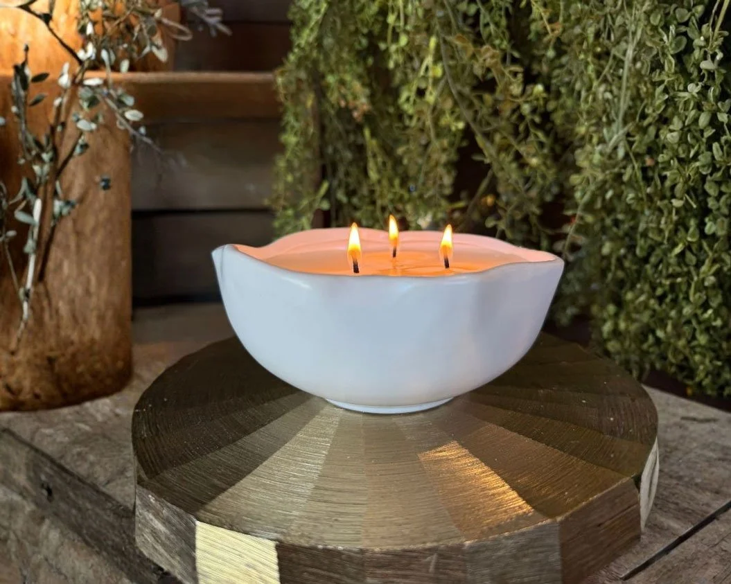 White ceramic bowl with three lit candles on a wooden surface surrounded by greenery.