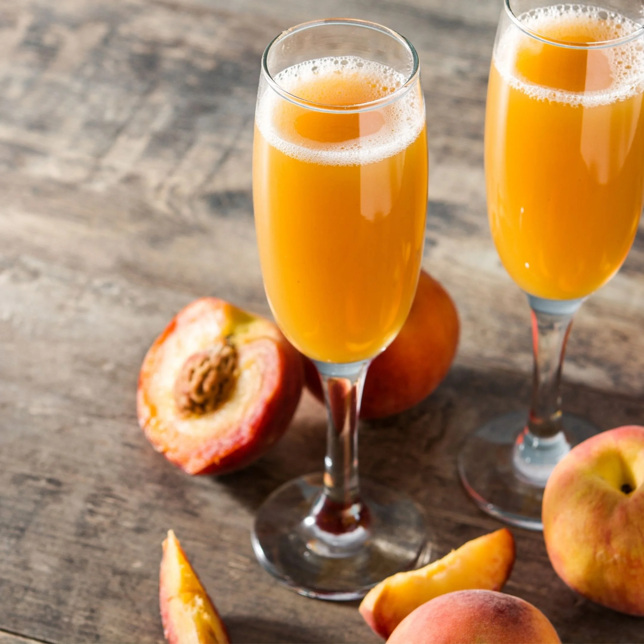 Two tall glasses of orange juice placed on a wooden surface surrounded by peeled peaches and peach slices.