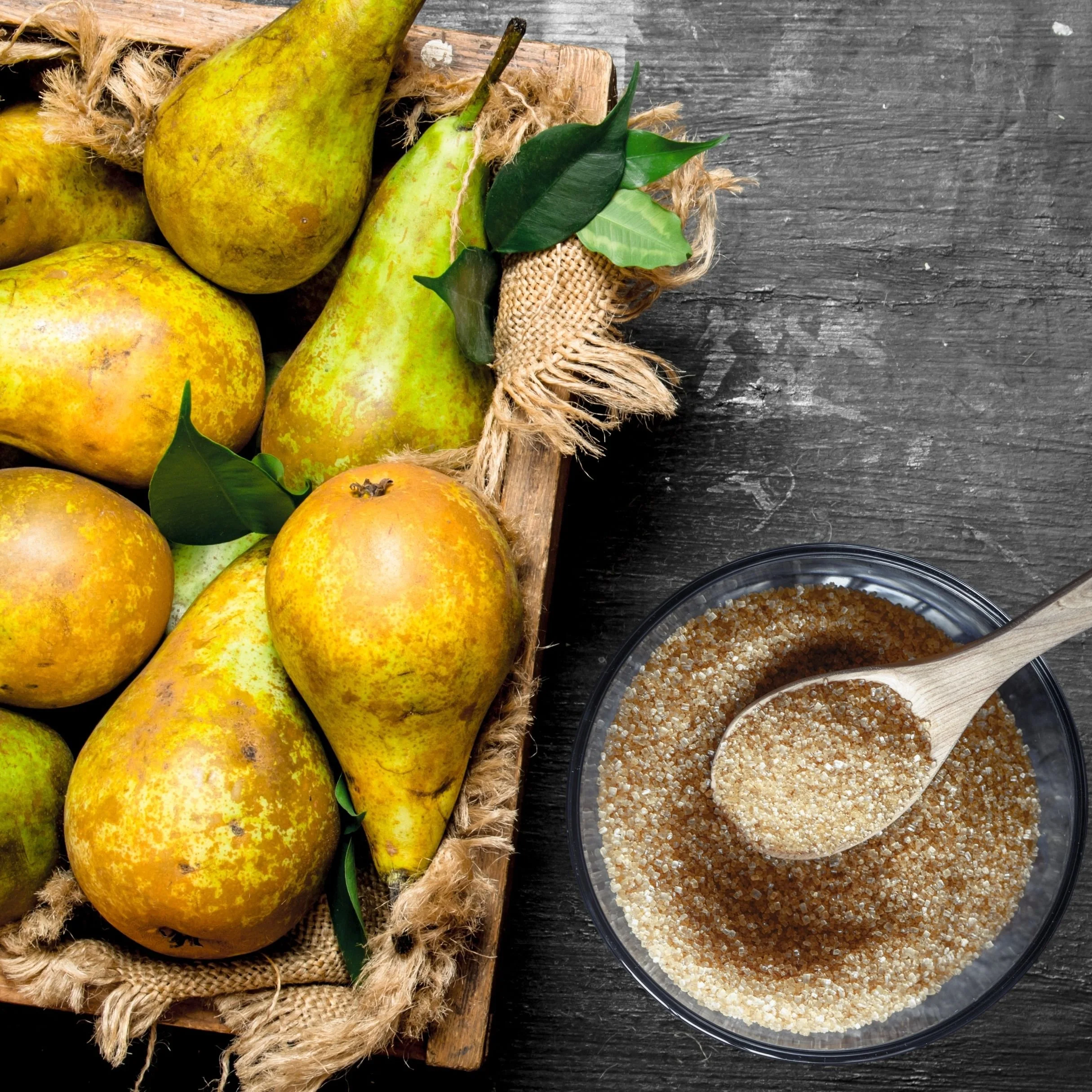 A wooden tray with yellow pears and green leaves, partially wrapped in burlap, on a dark wooden surface. Next to the tray is a glass bowl filled with brown sugar and a wooden spoon.