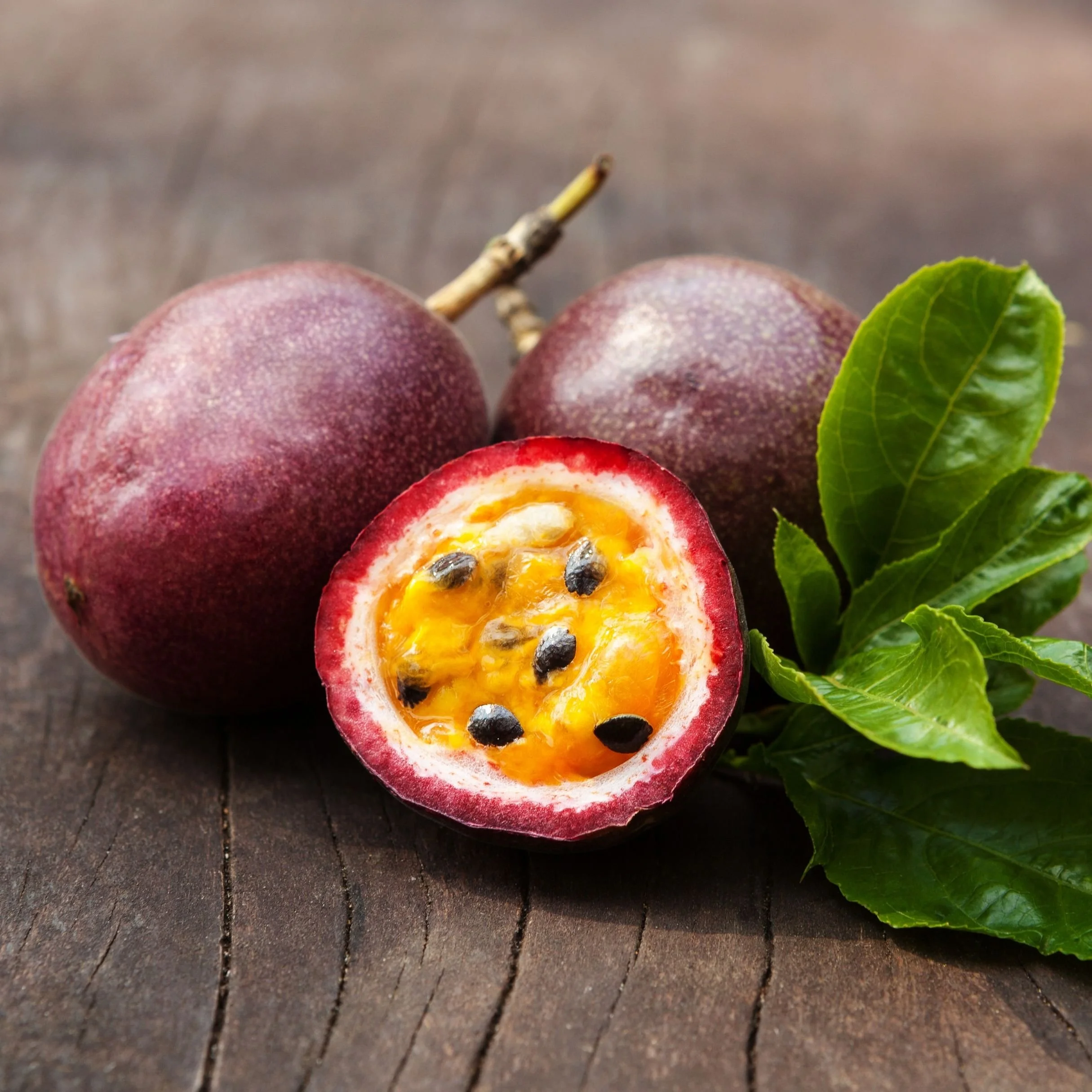 Close-up of a passion fruit with purple skin, cut open to reveal yellow pulp with black seeds, on a dark wooden surface with green leaves nearby.