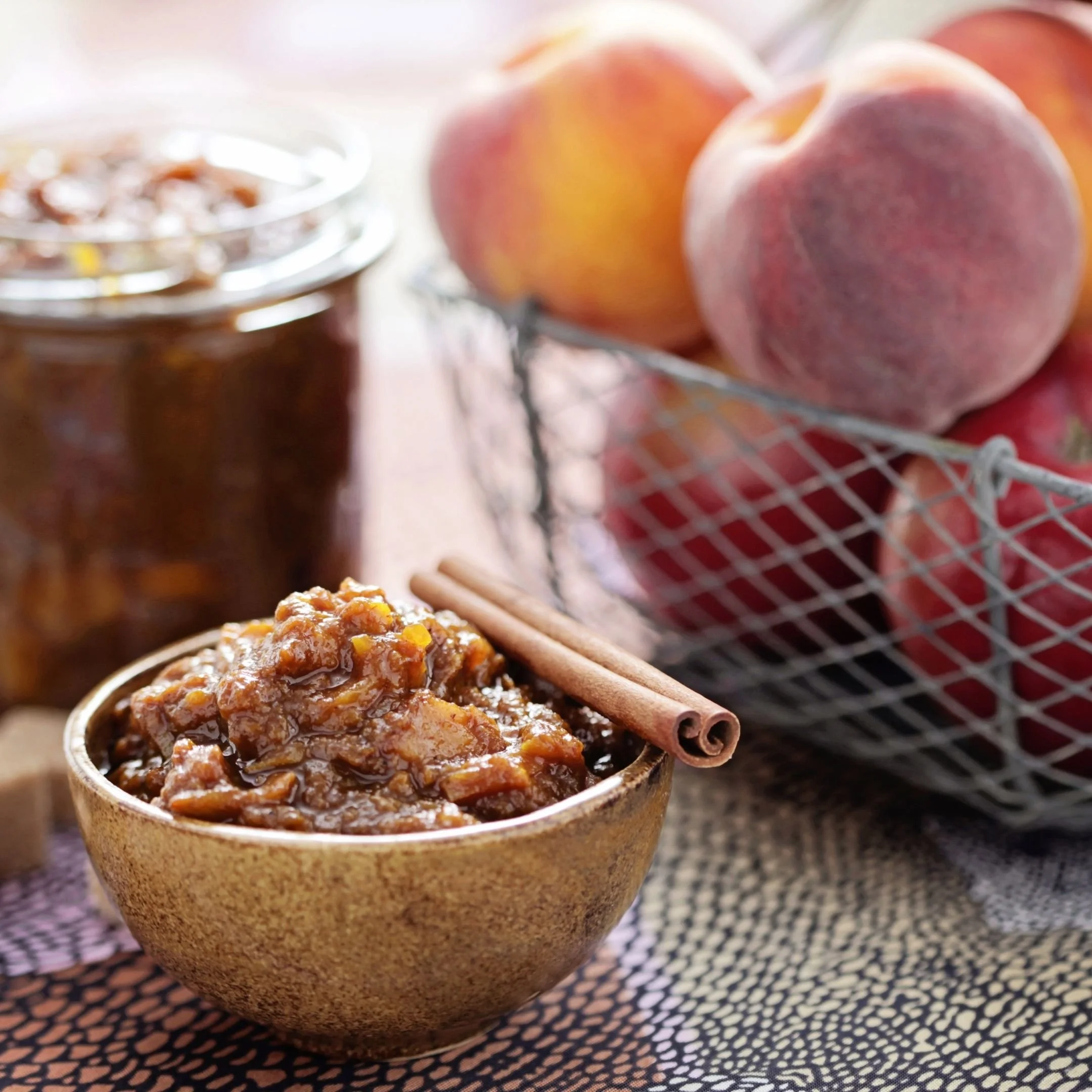 Close-up of a bowl filled with cooked chili, with a basket of peaches and cinnamon sticks in the background on a checkered cloth.