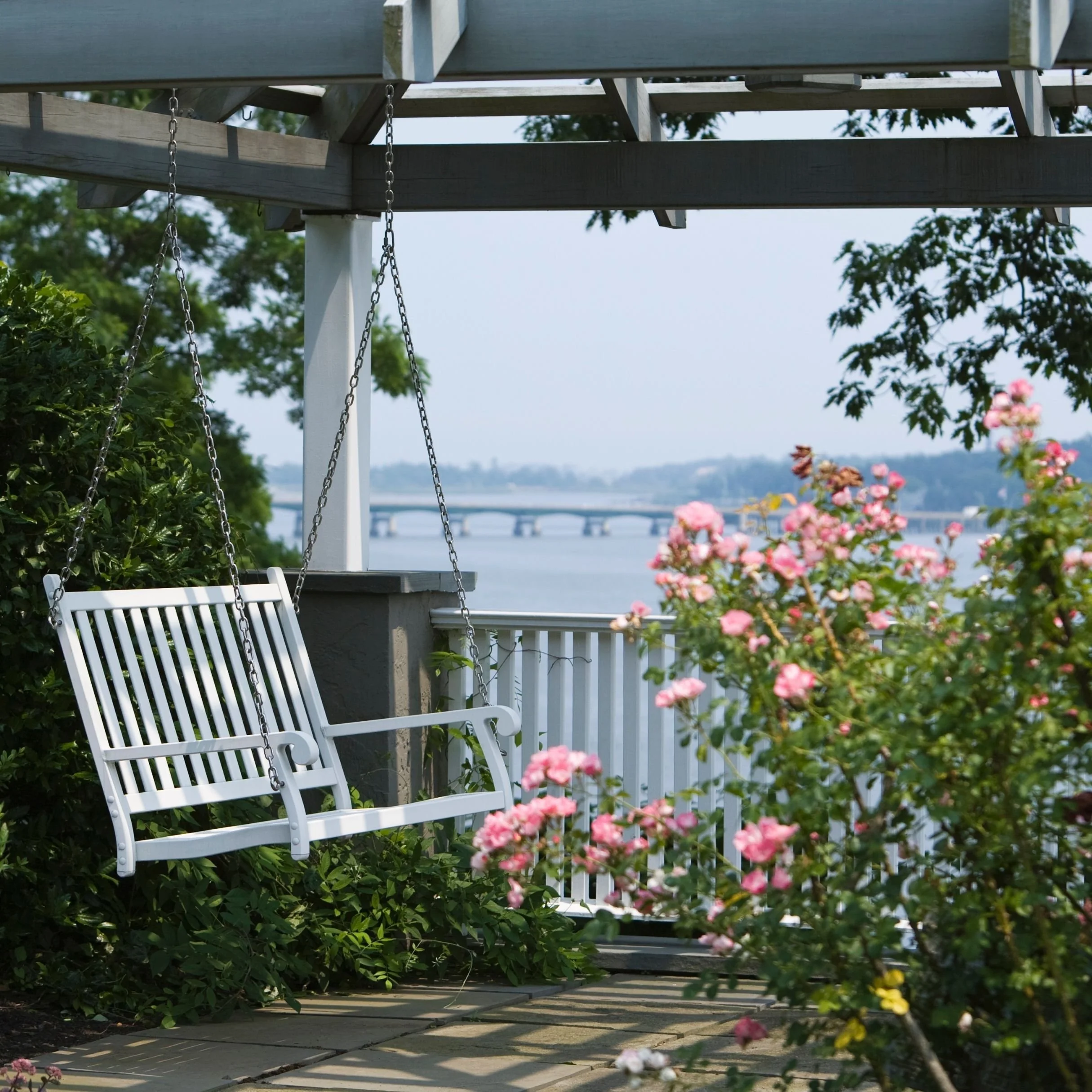 A backyard corner with a white porch swing hanging from a wooden deck, surrounded by greenery and pink flowers, overlooking a river and bridge in the background.