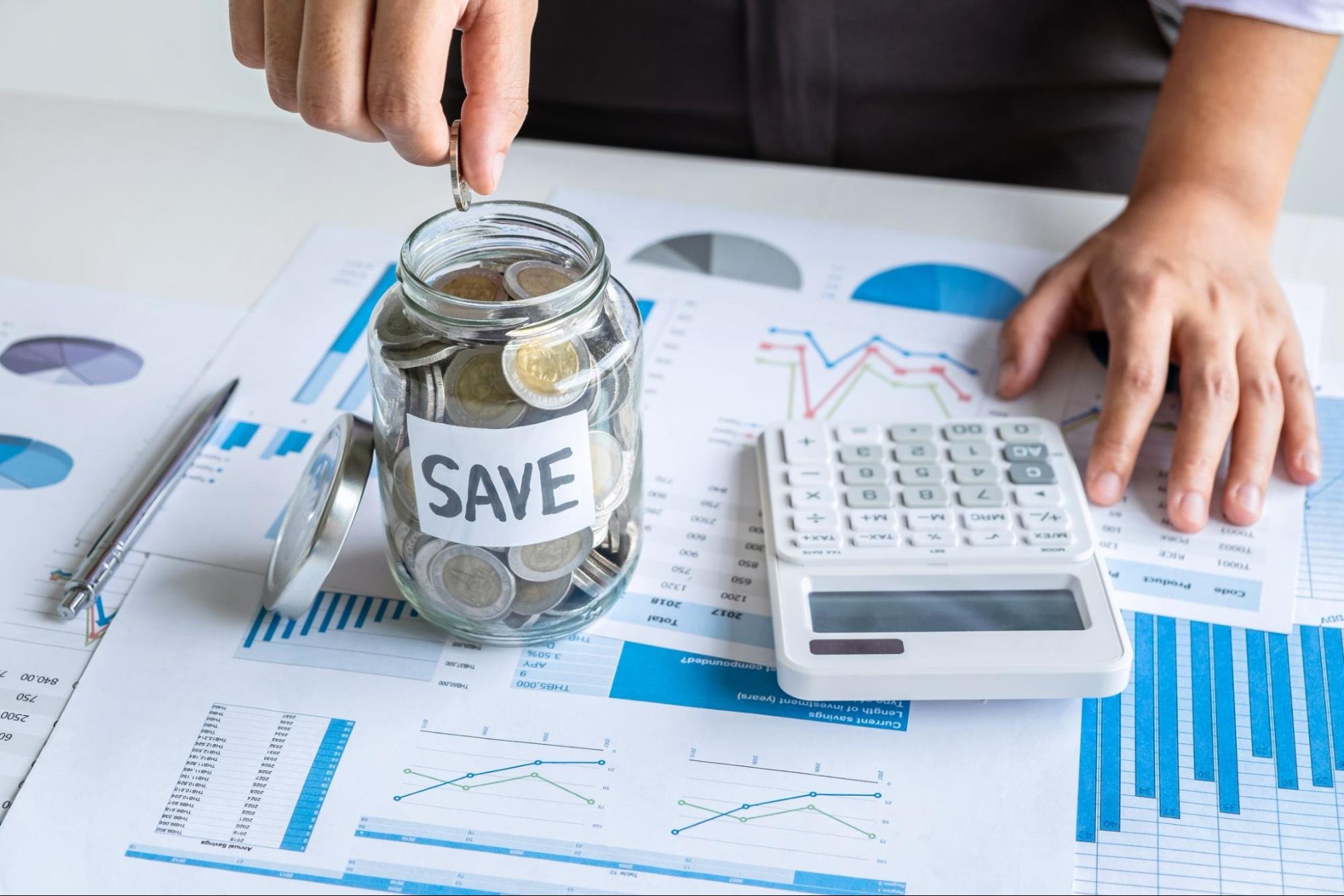Man putting a coin in a jar that says “save” next to a calculator and data on paper.