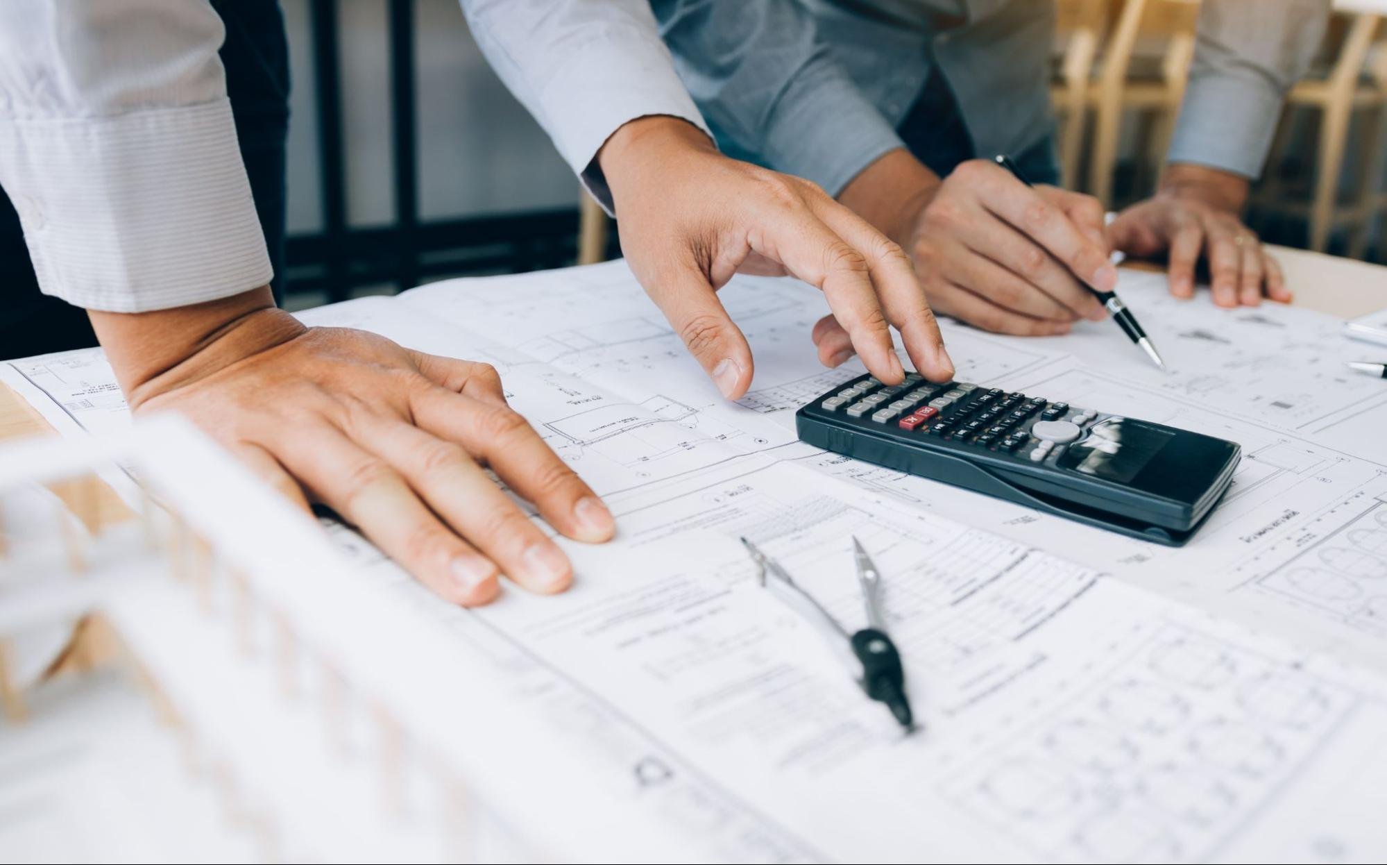 Professionals reviewing building survey costs and project plans with blueprints and a calculator in an office