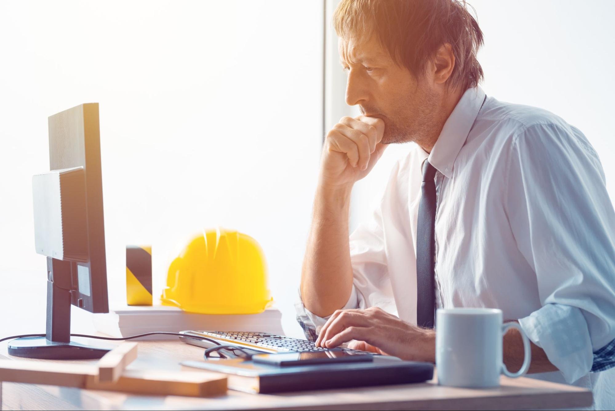A man sitting at his desk on his computer next to a hardhat for construction.