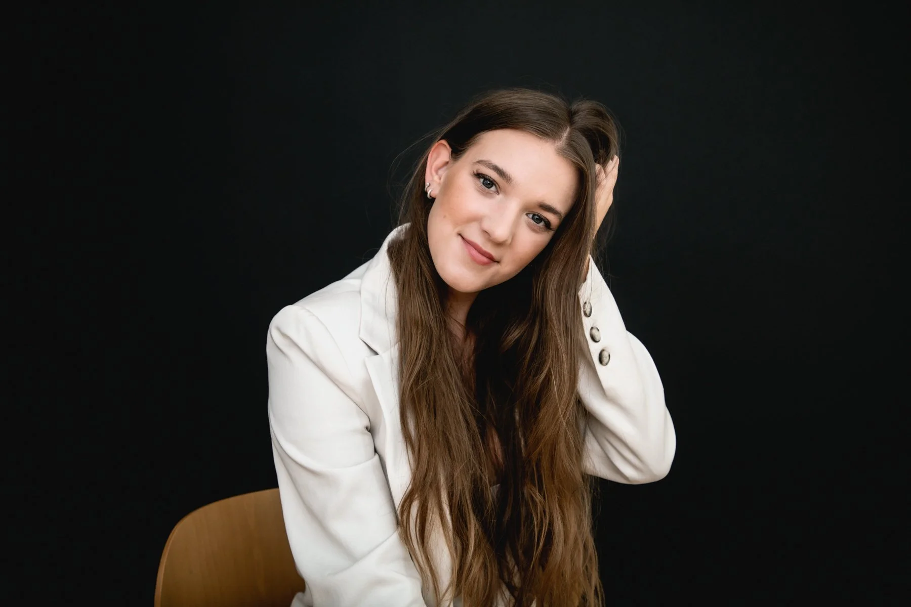 Portrait of a young woman with long brown hair, smiling and resting her head on her hand, sitting on a wooden chair against a black background.
