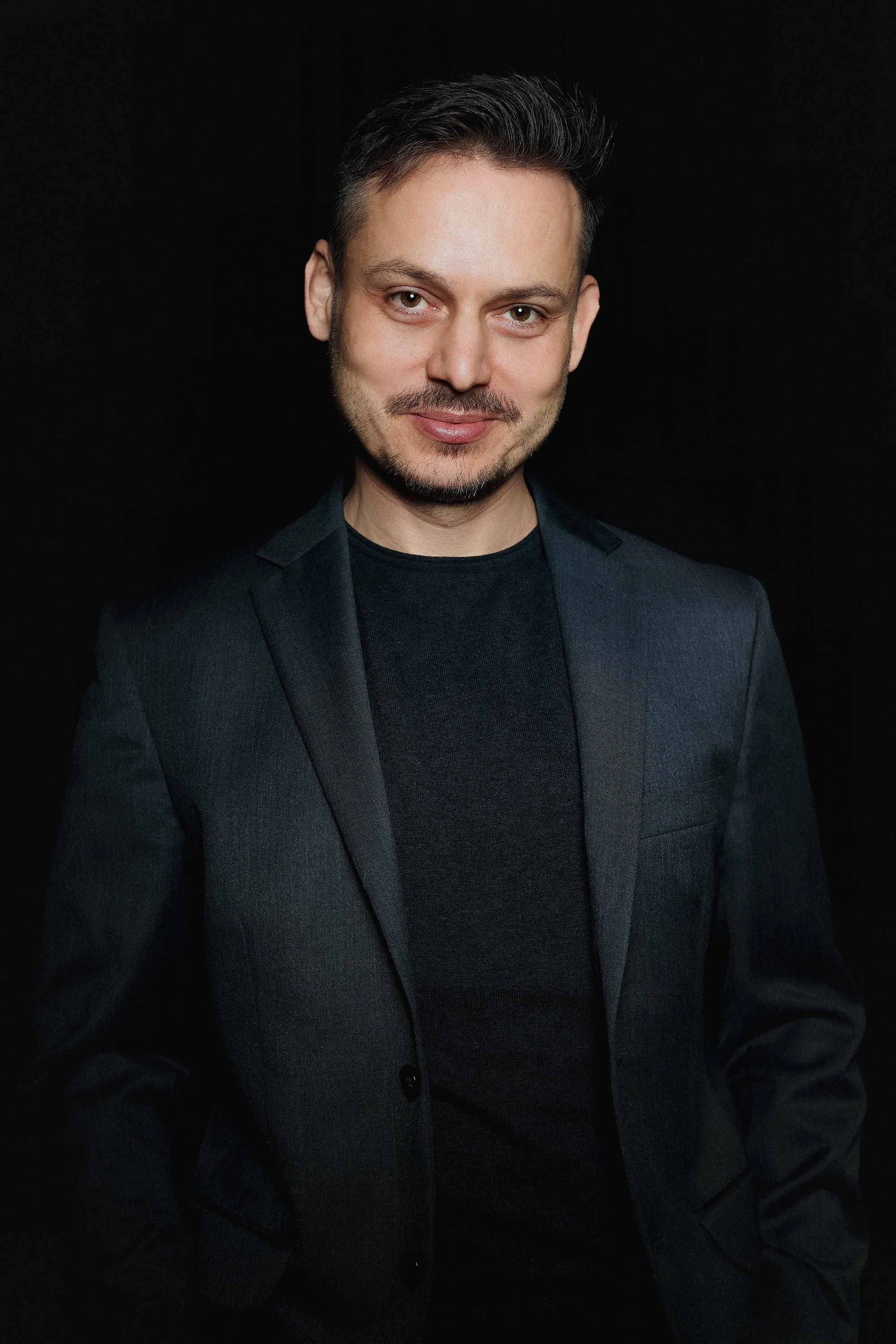 A man with short, dark hair and a beard, wearing a dark blazer and a black shirt, smiling slightly against a black background.