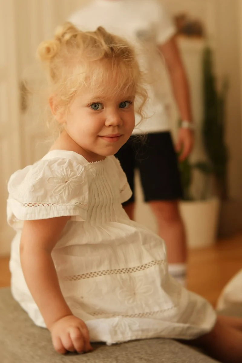 A young girl with curly blonde hair styled in small buns, wearing a white dress with embroidered flowers, sitting on a piece of furniture indoors.