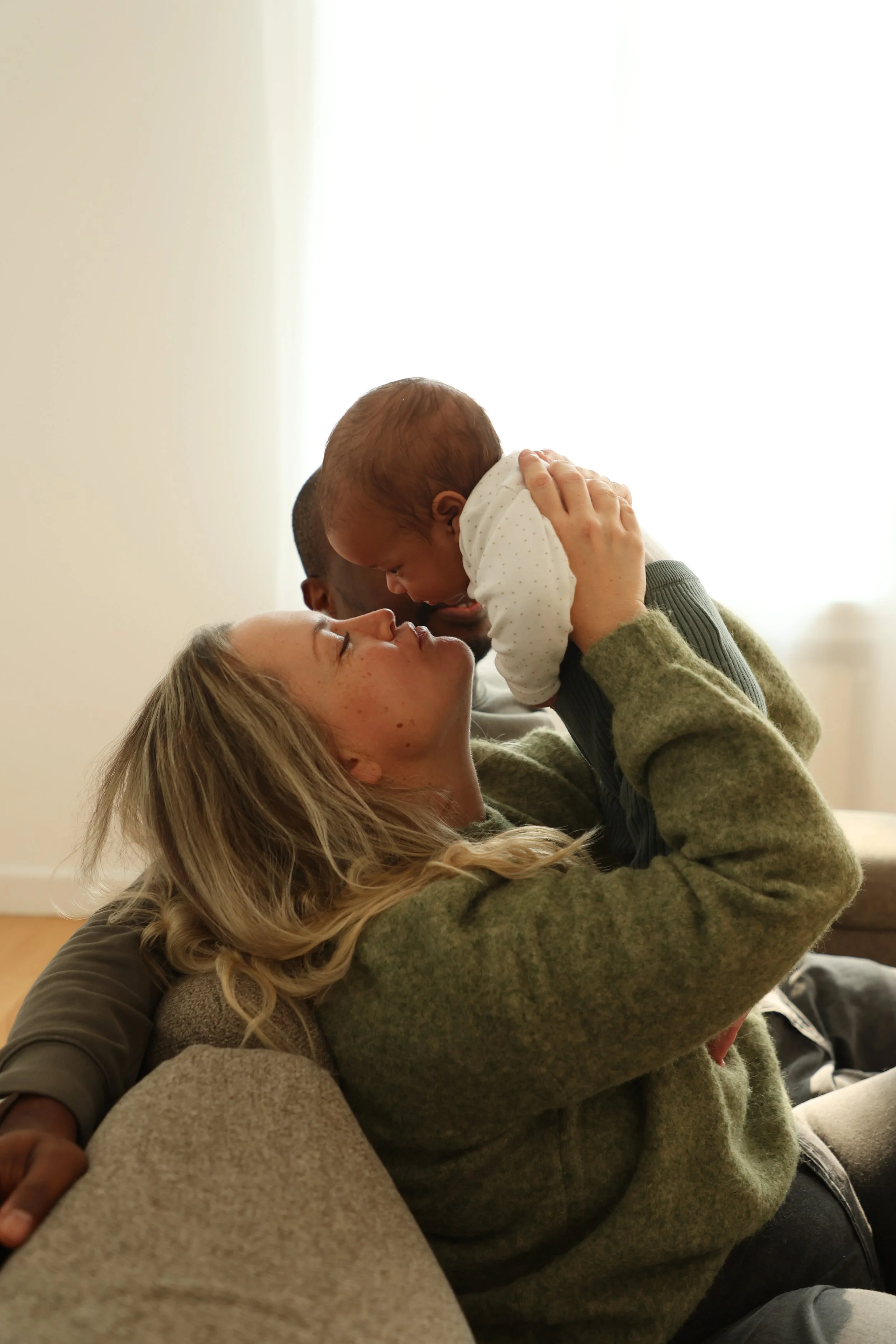 A woman holding a baby up in the air, both smiling and leaning toward each other indoors, with soft natural light streaming through a window in the background.