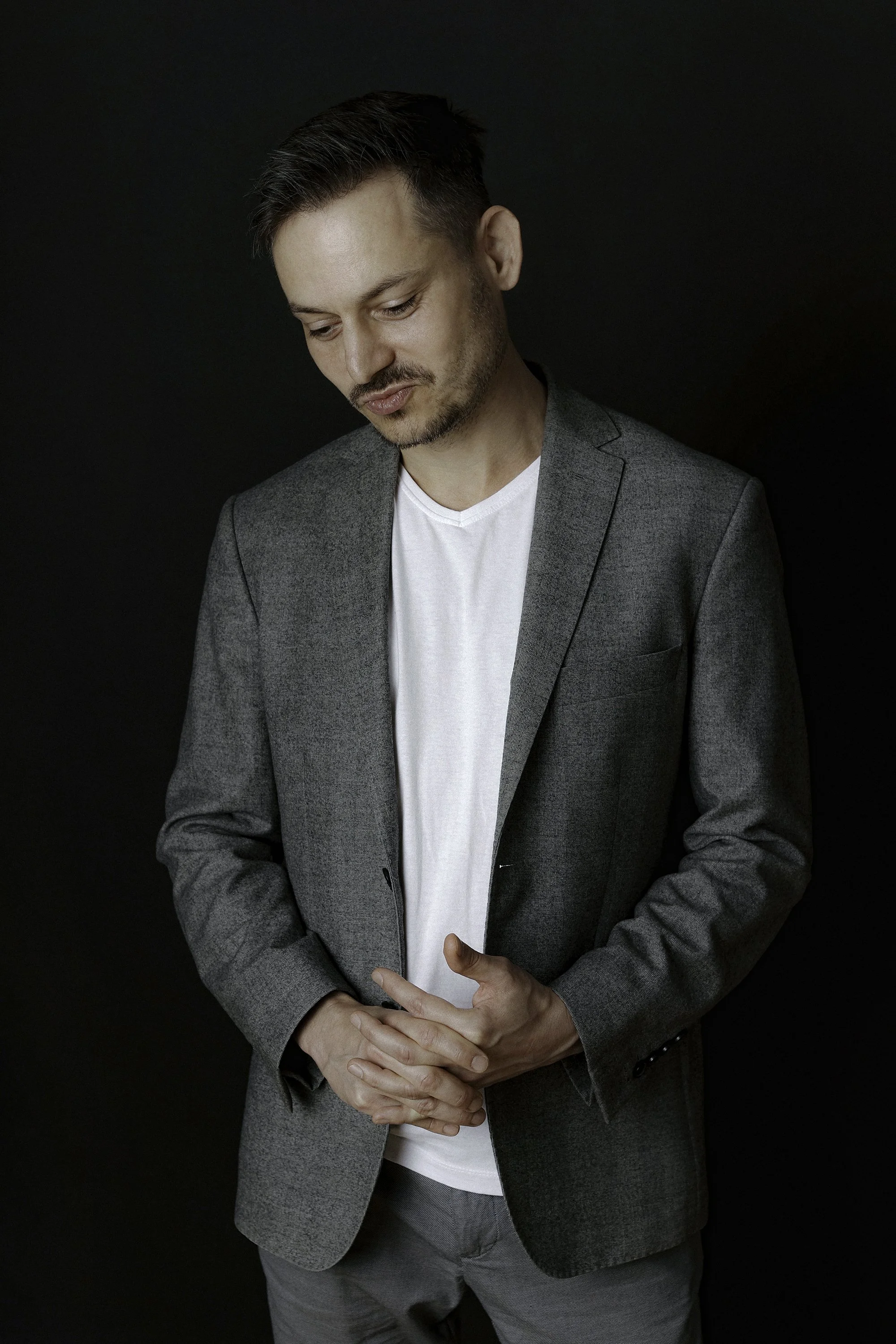 A man in a gray suit with a white T-shirt underneath, standing against a dark background. He has short, dark hair and facial hair, and is looking down with his hands clasped in front of him.