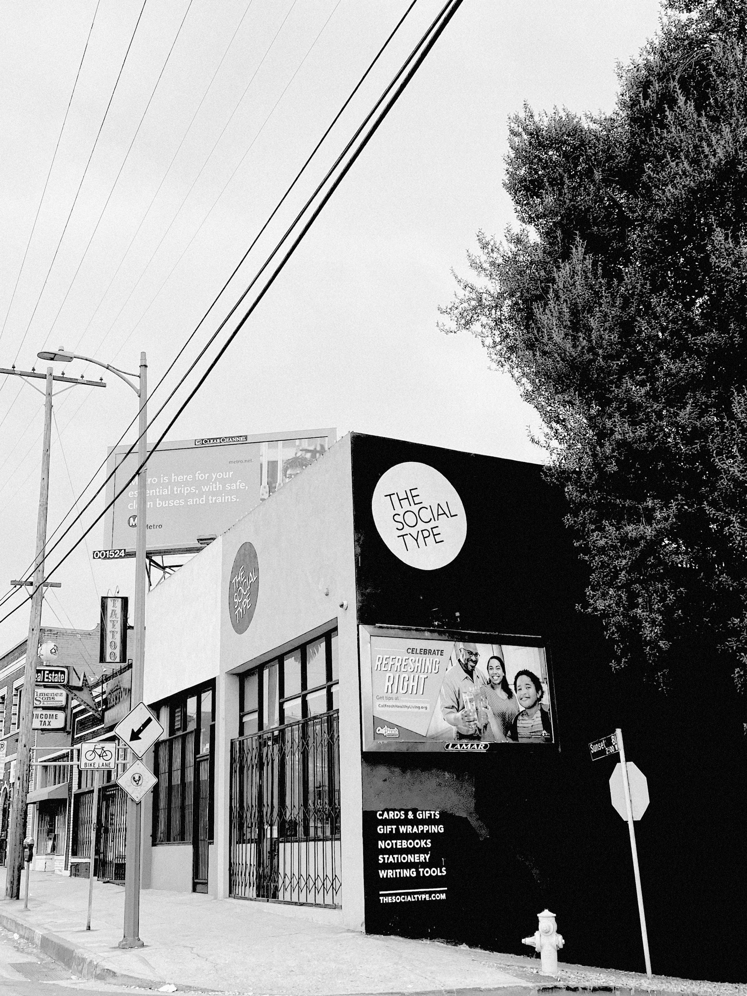 Black-and-white photo of a storefront with the sign 'The Social Type' and a billboard that says 'Celebrate Refreshing Right' with an image of three people. There are street signs and a fire hydrant nearby.