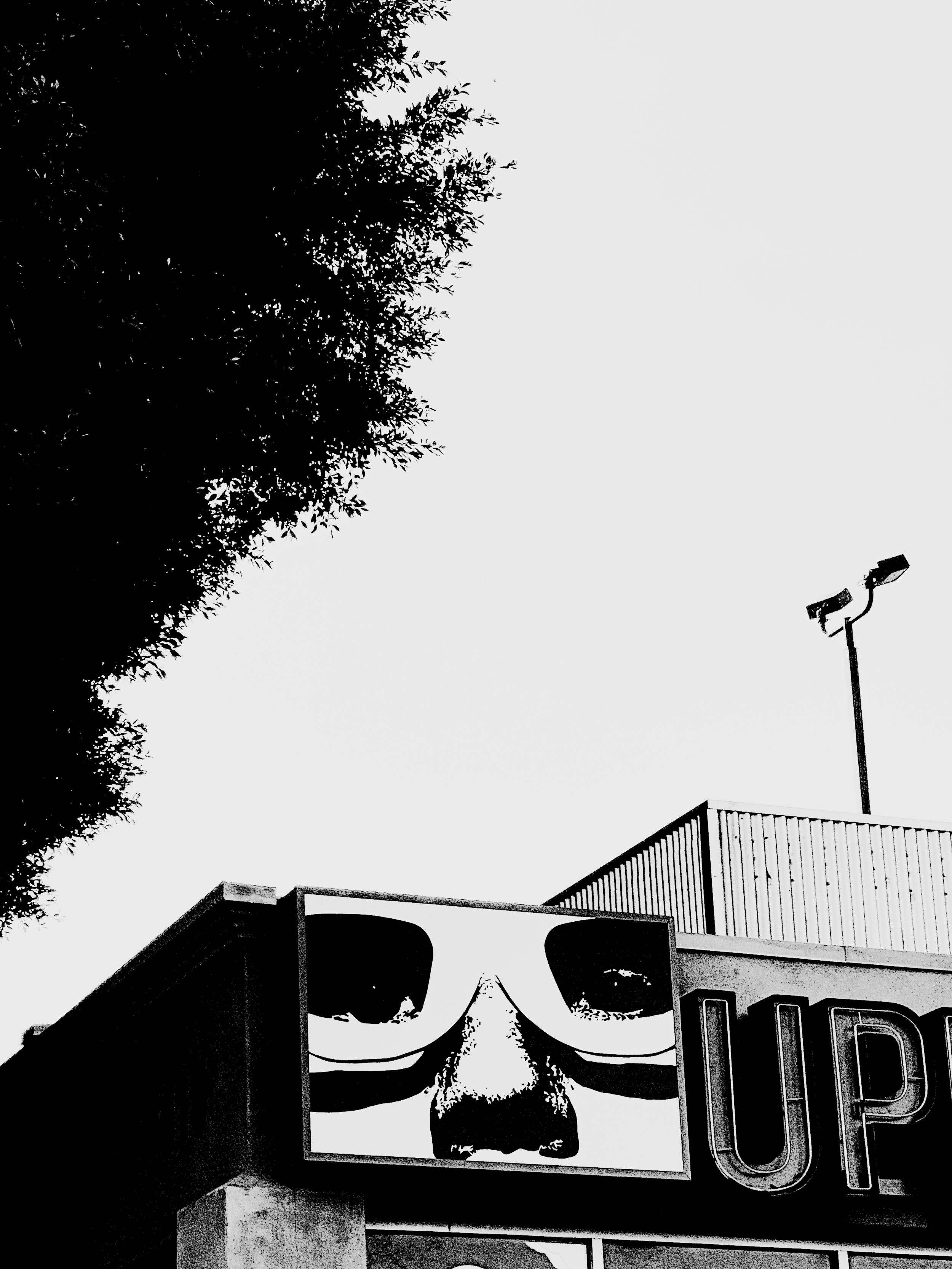 Sign with a face wearing sunglasses, showing only the eyes and nose, attached to a building with the partially visible word 'UP' in large letters.