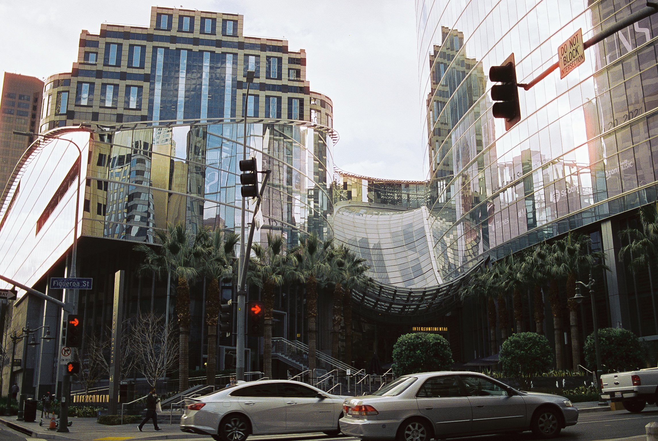 Modern glass building with curved architecture, palm trees, traffic lights, and vehicles on the street in front.