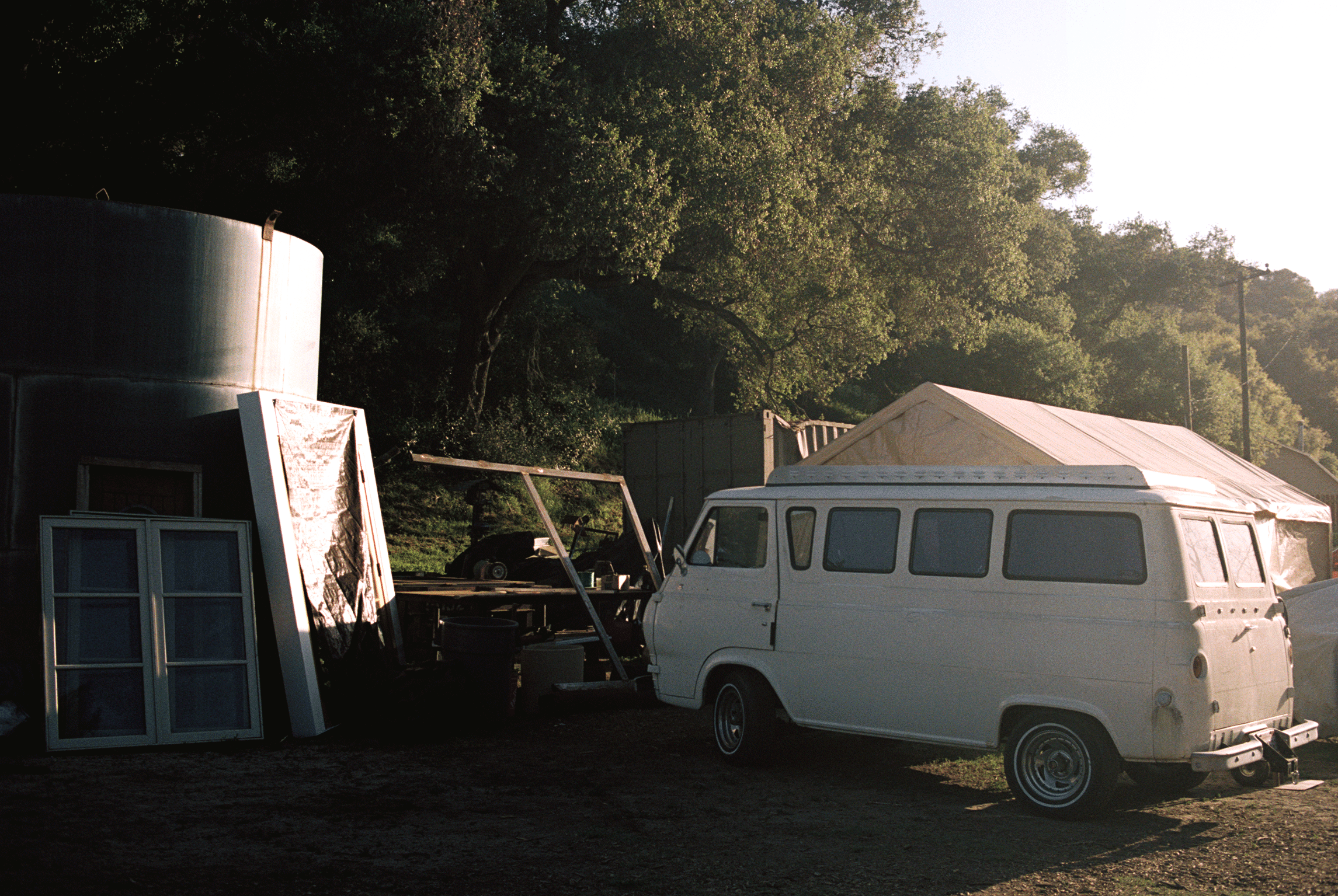 An outdoor scene with a white vintage van parked on the ground, a tent, and a shed or trailer, with trees and sunlight in the background.