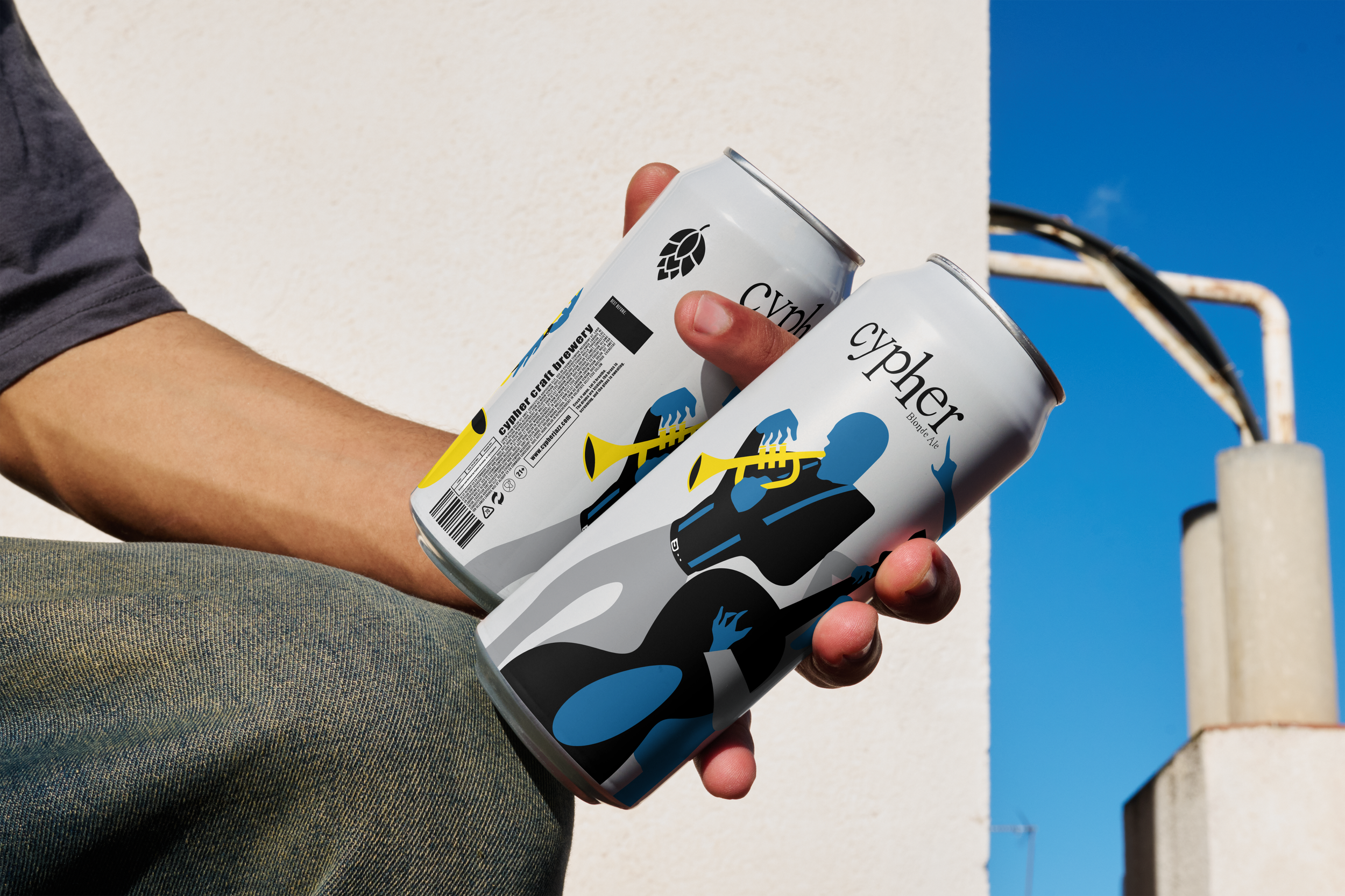 Person holding two cans of Cypher Blonde Ale outdoors with a blue sky and white building in the background.
