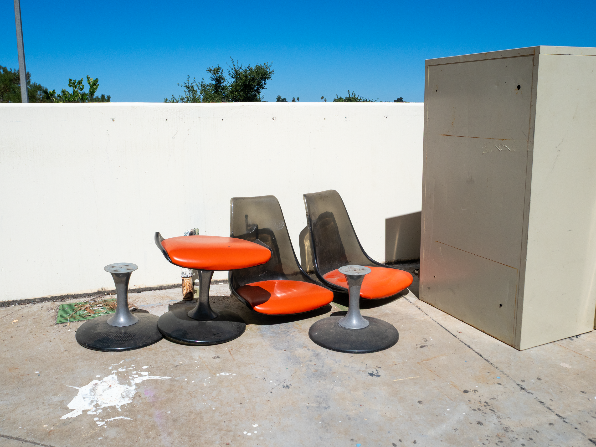 An outdoor scene with three red and black chairs, a white wall, a beige storage cabinet, and some green plants and trees in the background under a clear blue sky.