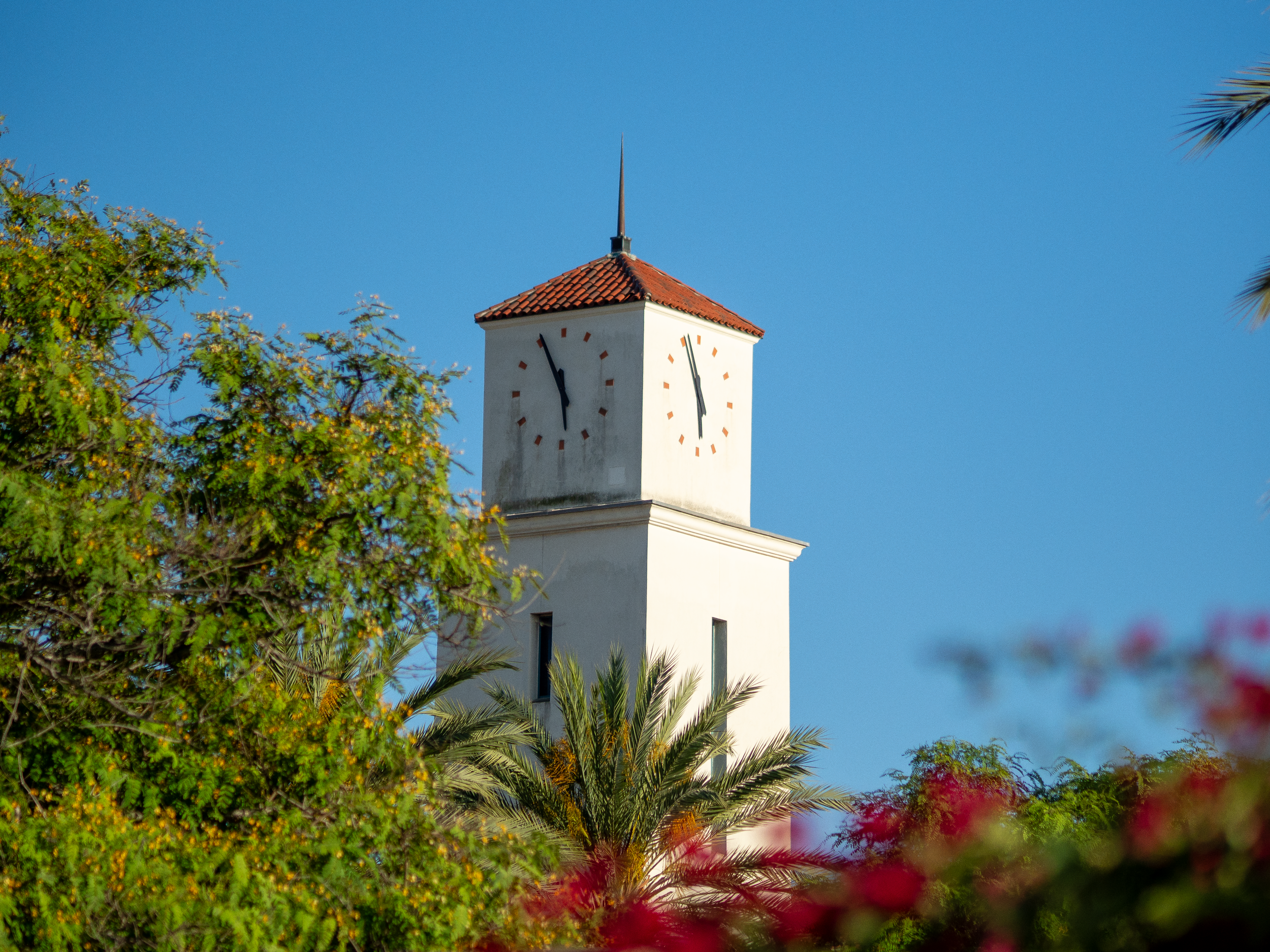 A white clock tower with a red tiled roof and a sharp spire on top, surrounded by lush green trees and plants, set against a clear blue sky.