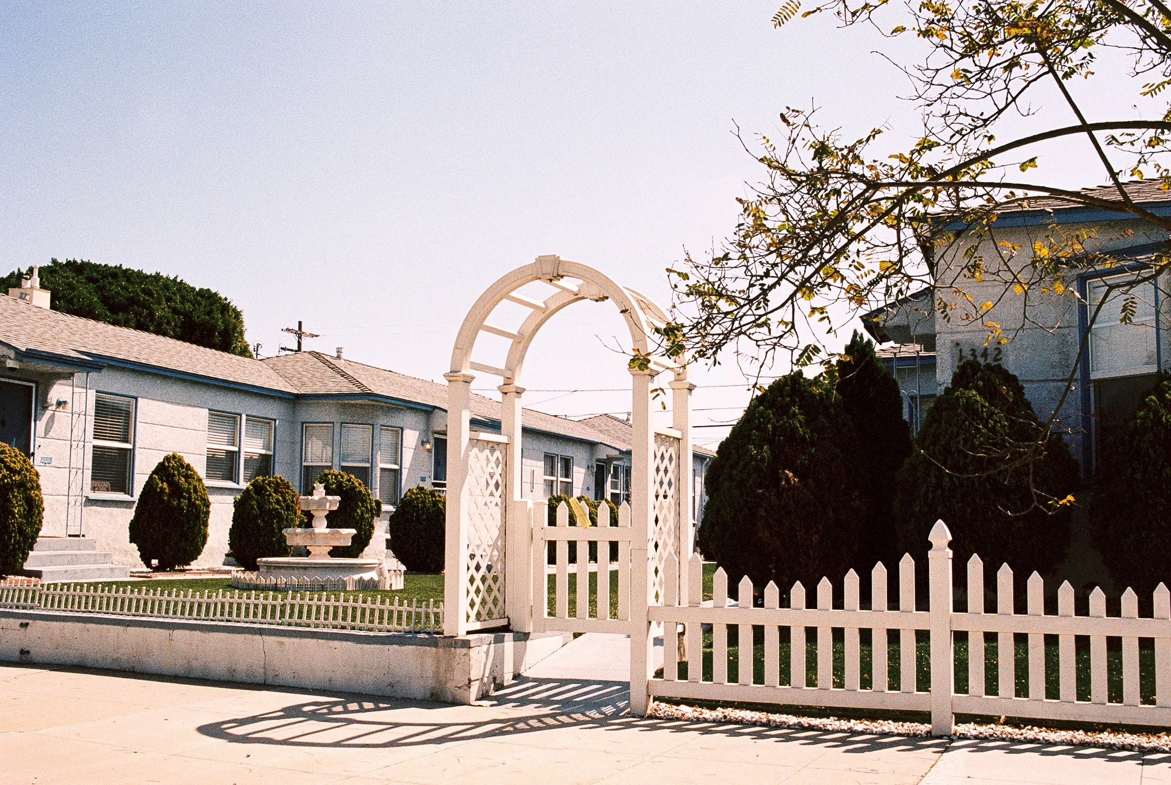 Front yard with white picket fence, white gate, stone fountain, trimmed bushes, residential buildings, and a blue sky with trees.