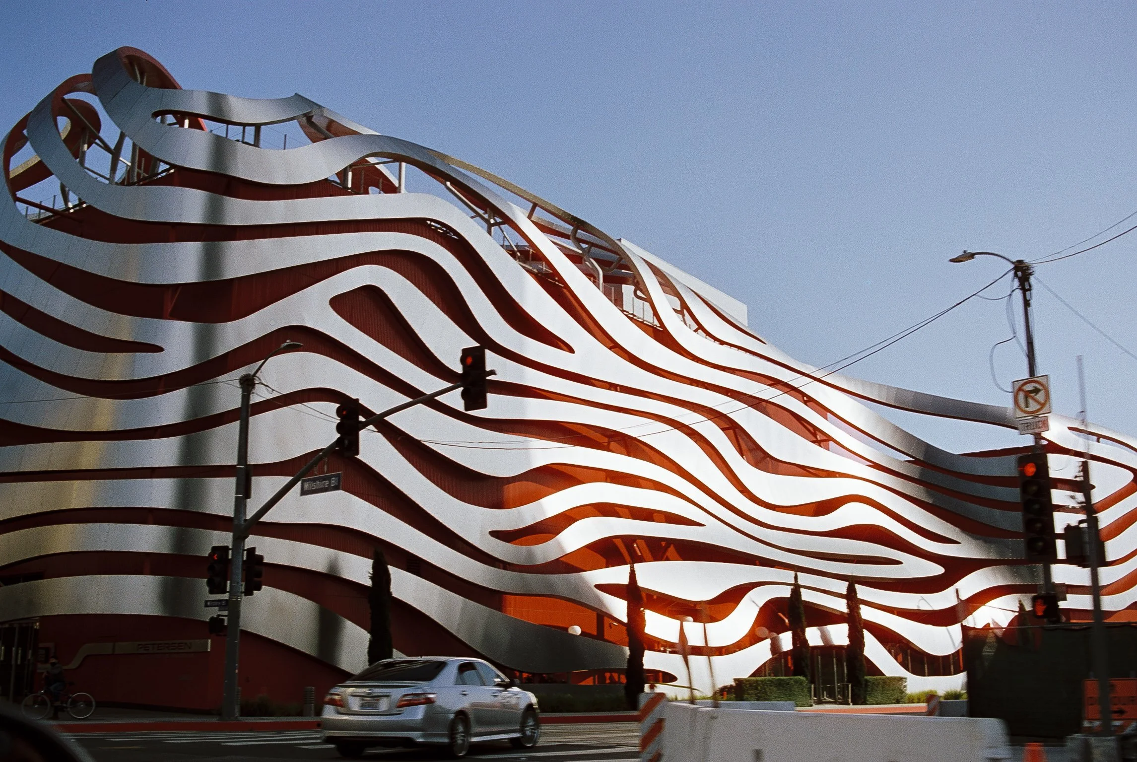 Modern building with wavy, layered design in silver and red, with streetlights, traffic signals, cars, and a clear sky visible.