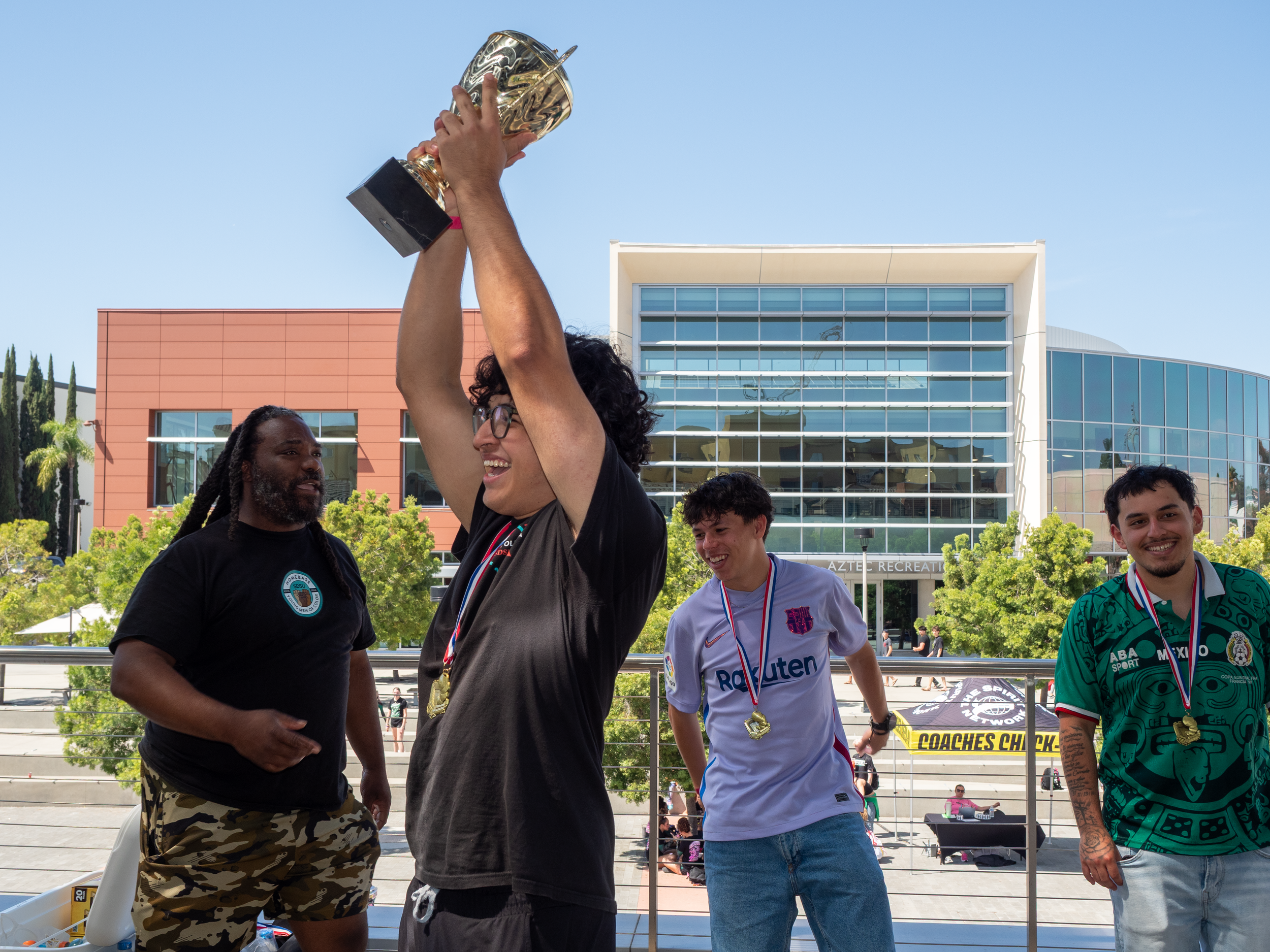 Group of people celebrating with medals, one person holding a trophy up in the air, outside in front of modern buildings and trees.