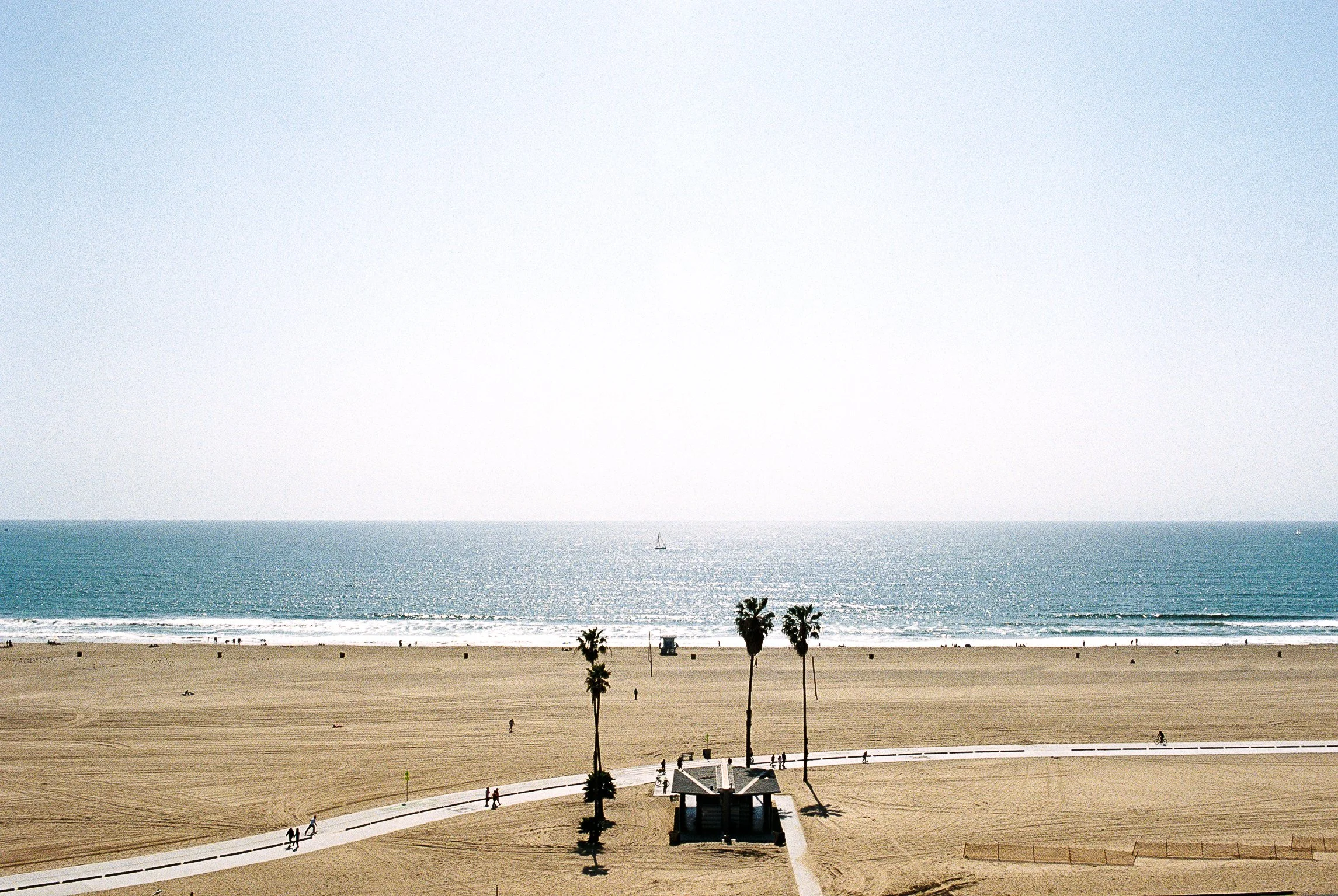 A sandy beach with a few palm trees and people walking along the promenade. The ocean is calm, and there's a boat in the distance under a bright, clear sky.