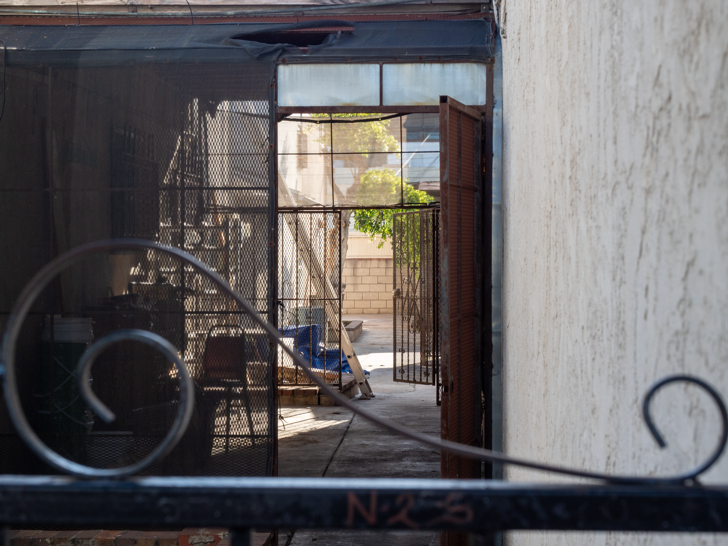 View through a metal gate leading from an interior space to an outdoor area with trees and buildings in the background, showing a construction or renovation site.