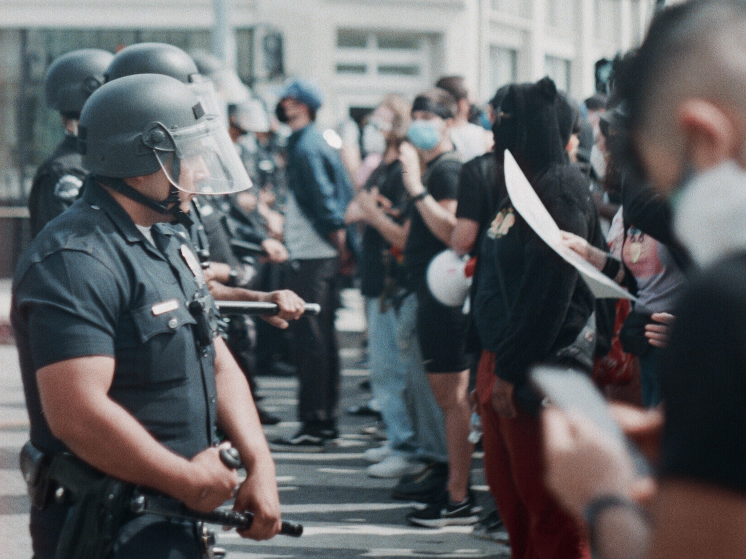 Police officers facing protesters on the street during a confrontation, some protesters holding signs and wearing masks.
