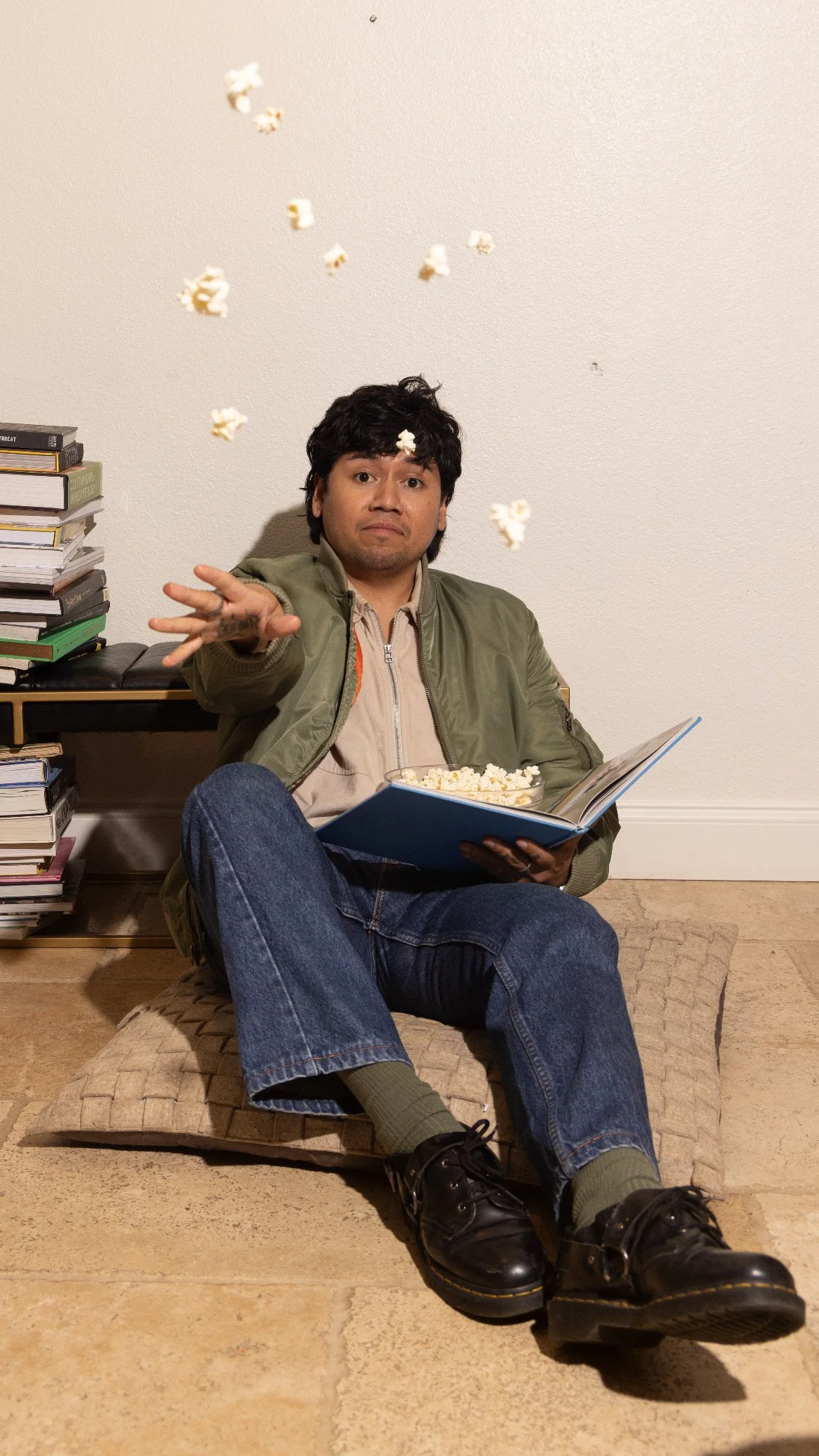 A man sitting on a cushion on the floor, holding a book with popcorn in his lap, with popcorn falling from above, against a plain white wall, with a stack of books on the left.