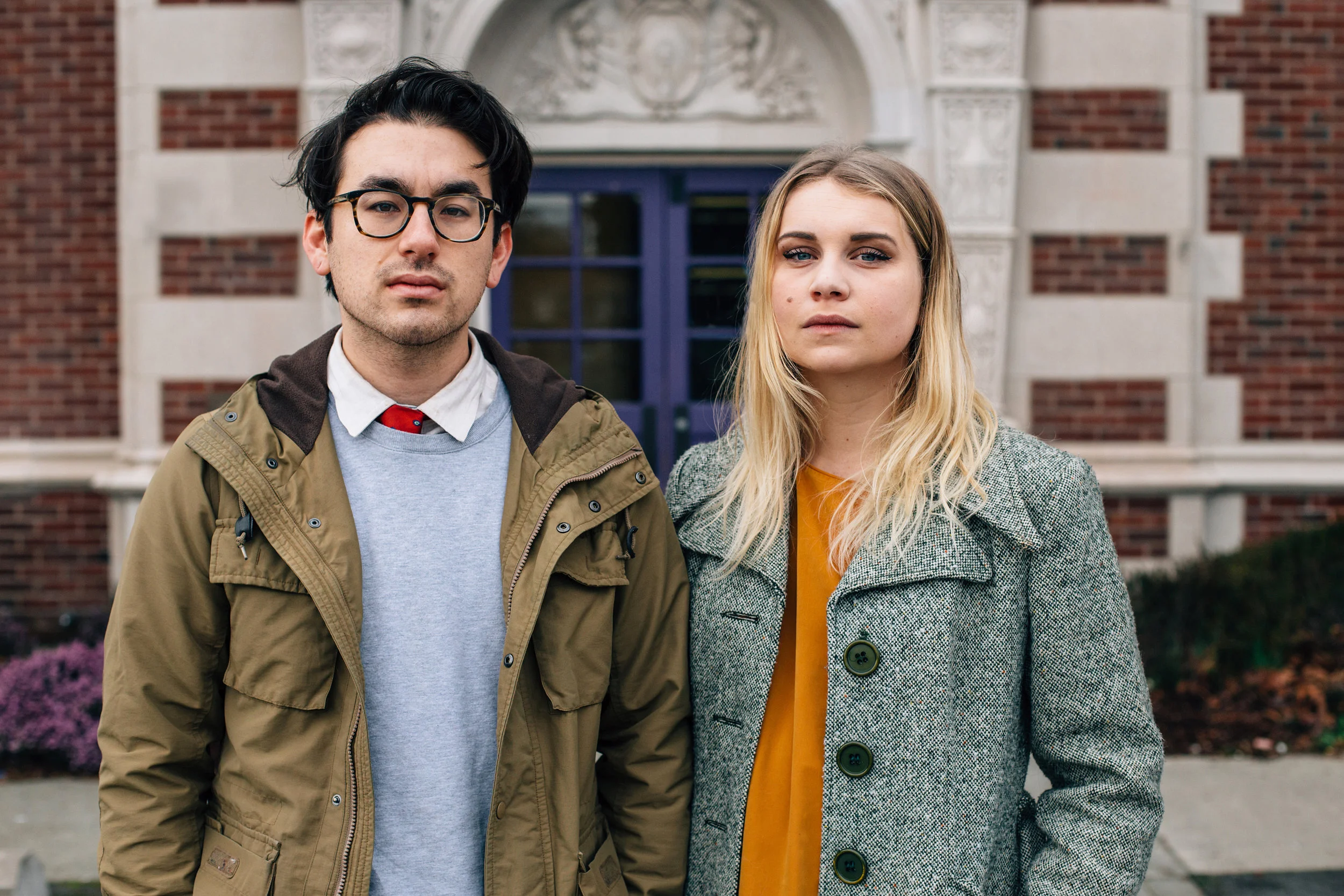 A young man and woman standing outside in front of a brick building with a purple door, looking directly at the camera with serious expressions.