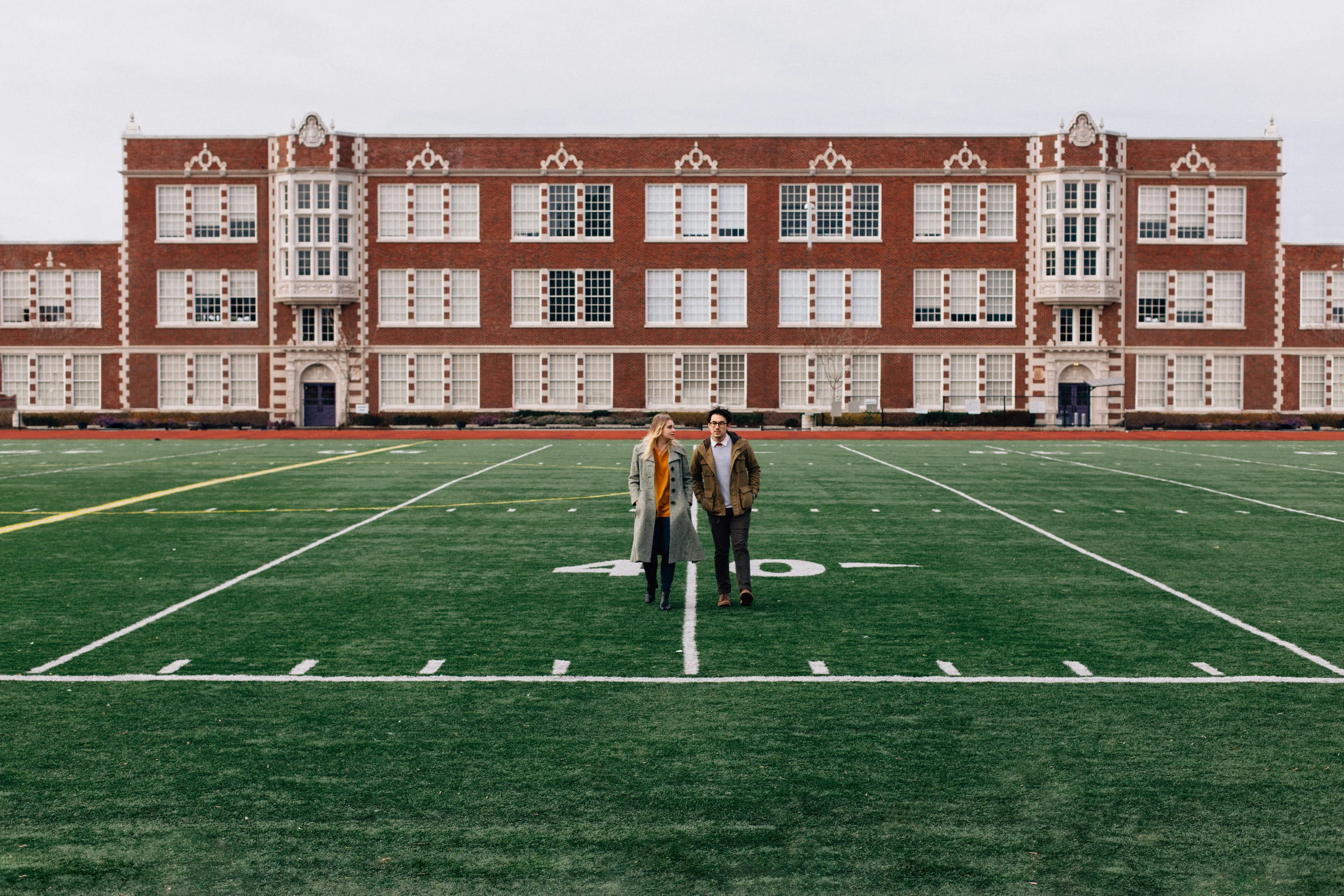 A man and woman walking on a grassy sports field with a large historic brick school building in the background.