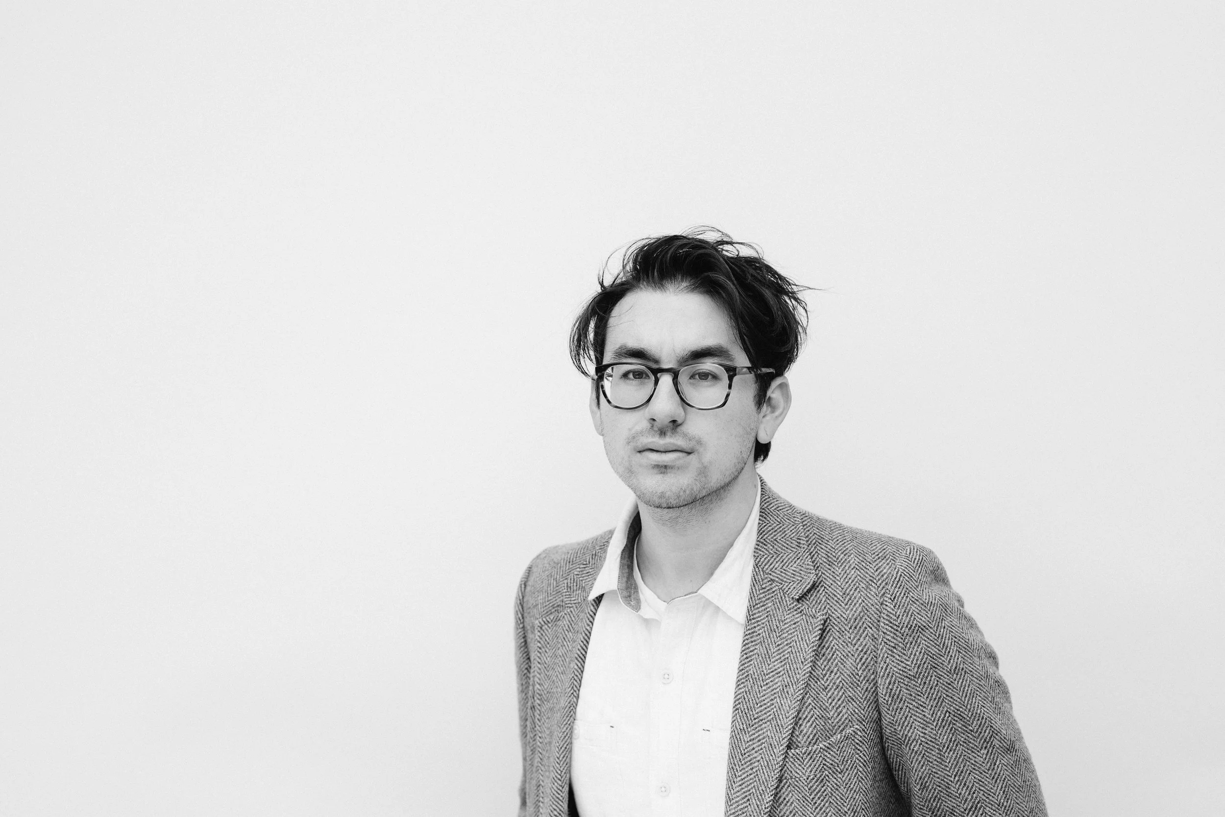 Black and white portrait of a young man wearing glasses, a checkered blazer, and a white shirt, standing against a plain background.