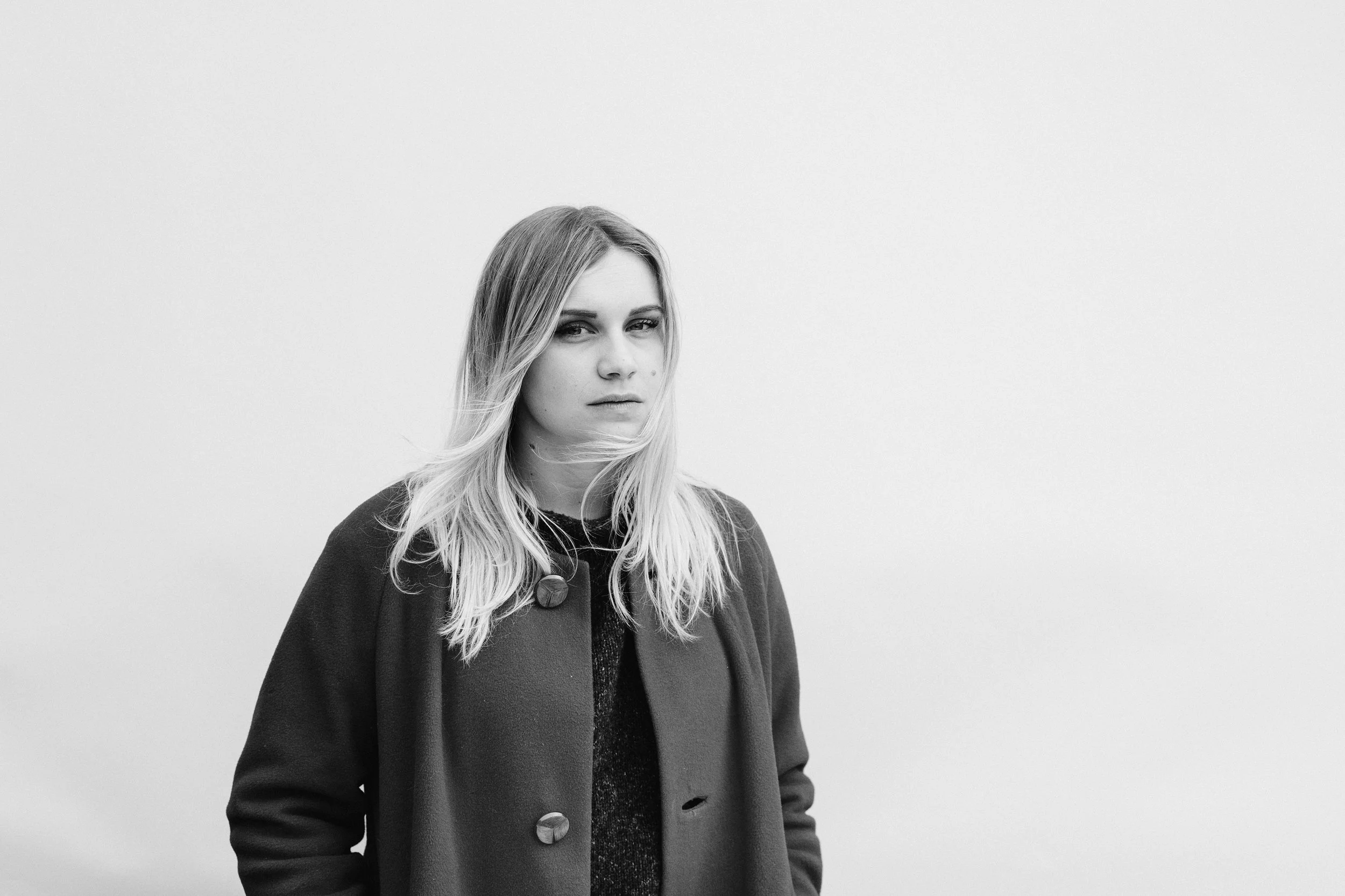 Black-and-white portrait of a woman with long blond hair, wearing a dark coat with large buttons, standing against a plain, light background.