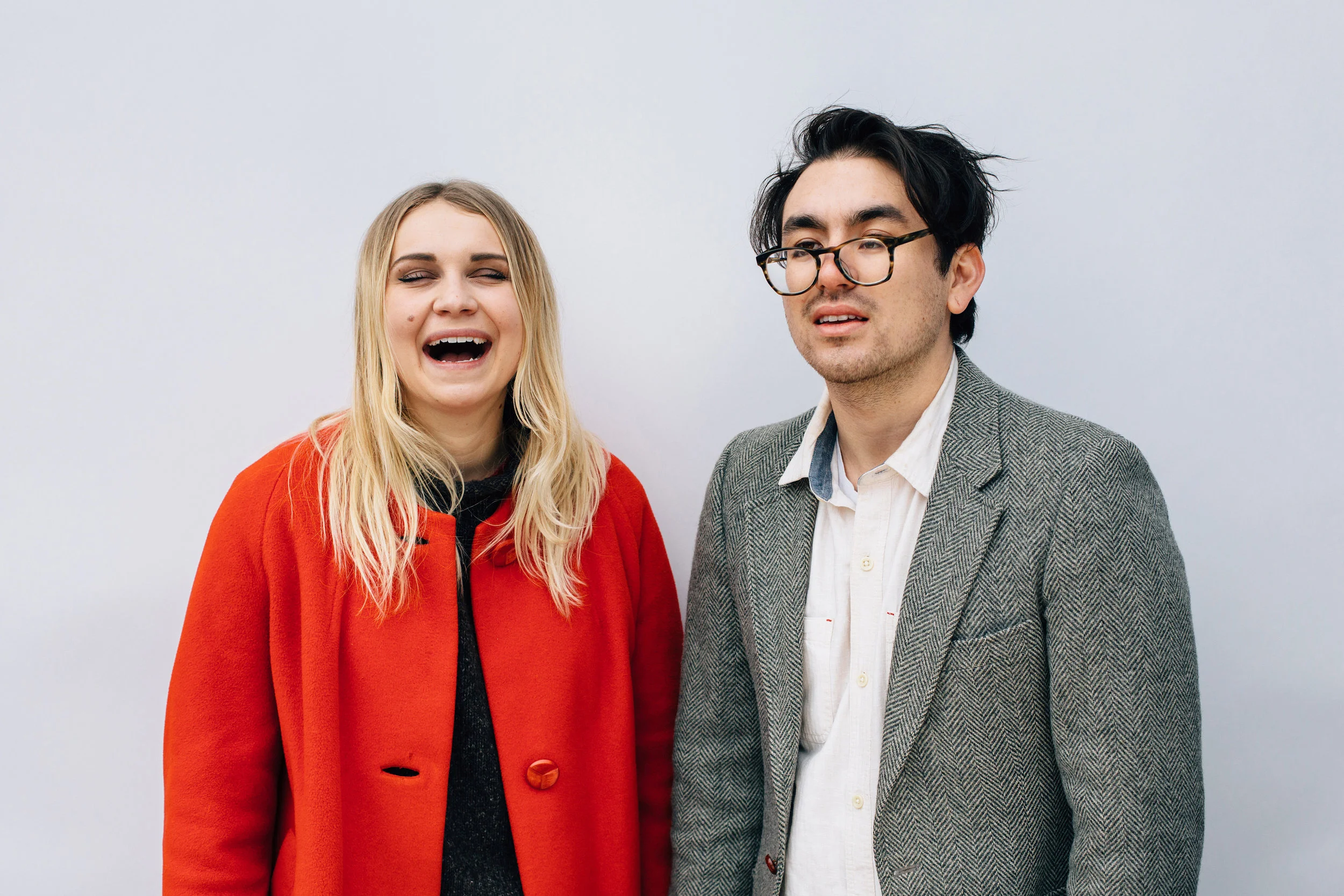 A woman with blonde hair smiling and a man with glasses and dark hair standing against a plain gray background.